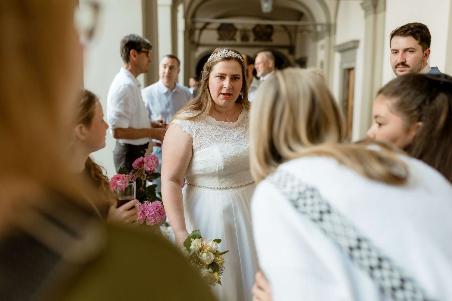 Bride in a white lace dress holding pink flowers while greeting guests at an elegant wedding reception.