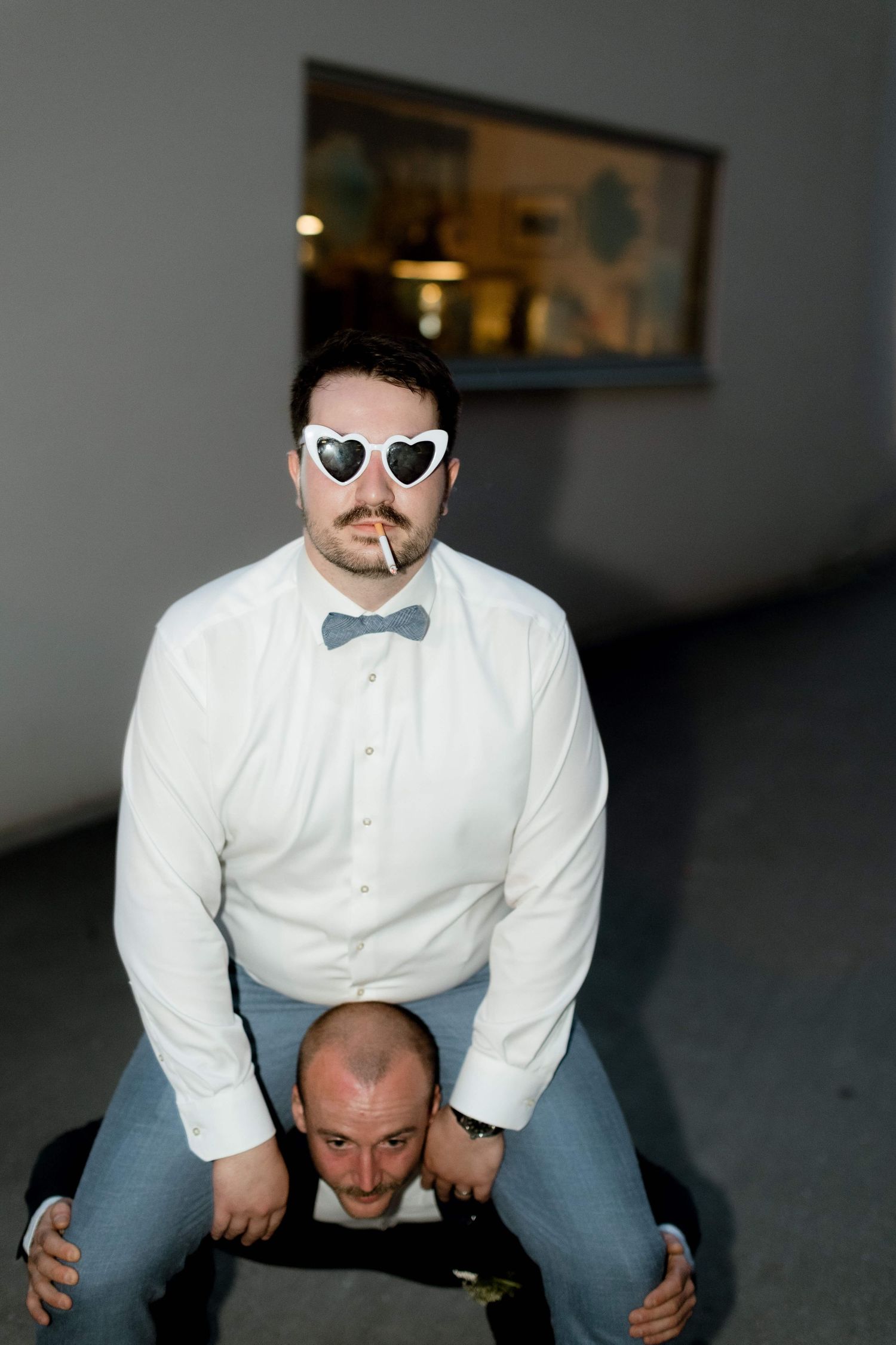 Man in heart sunglasses and bow tie sitting on another mans shoulders indoors.