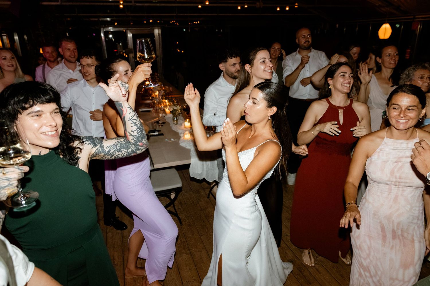 Guests and bride in white dress dancing energetically at lively indoor wedding reception.