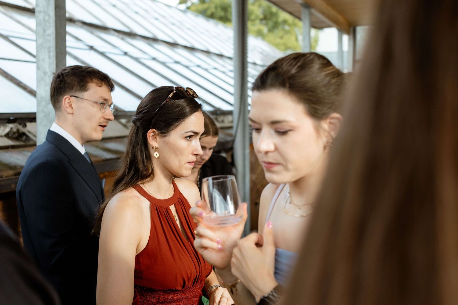Two women in elegant dresses conversing over drinks at an indoor wedding reception in a greenhouse venue.