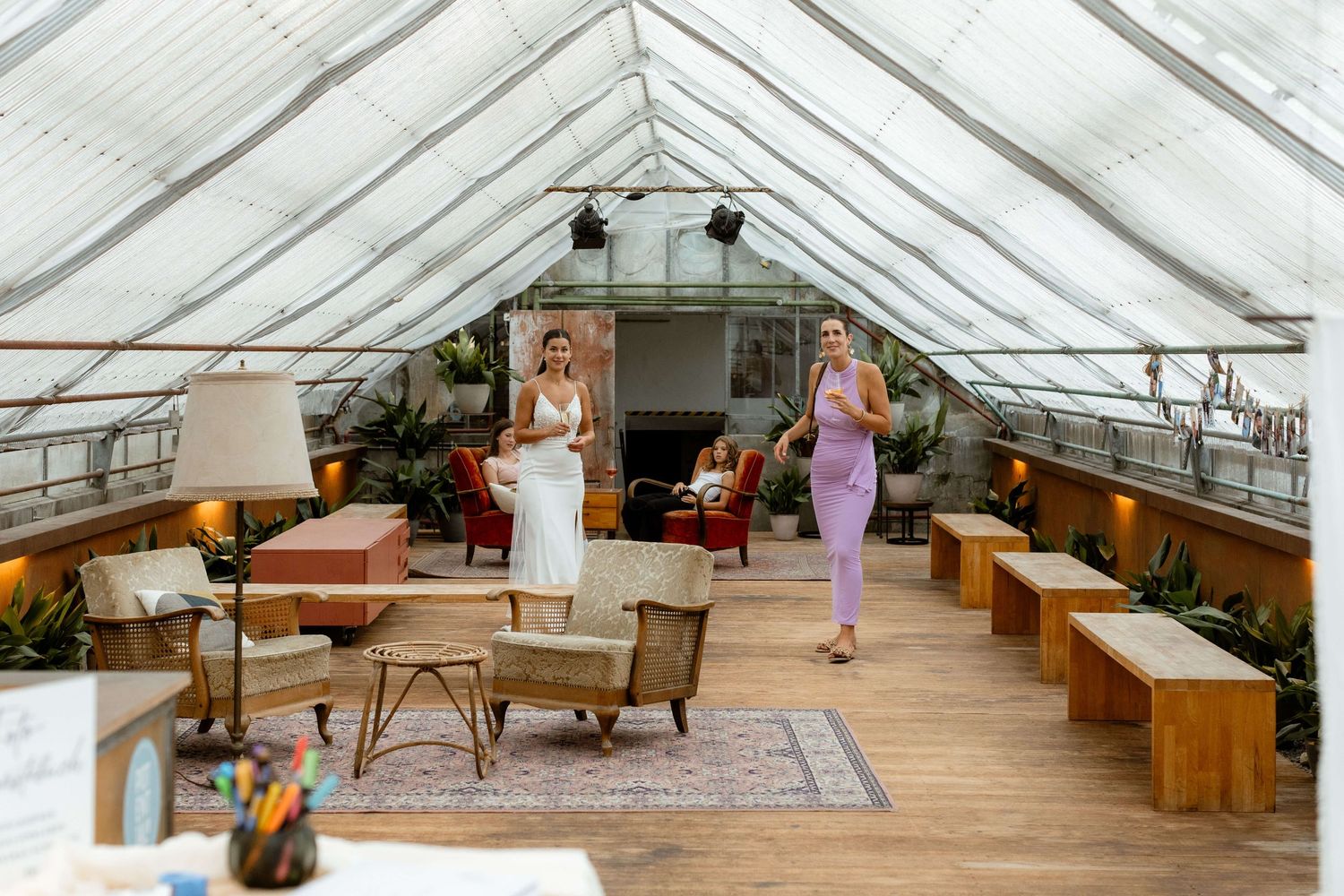 Two women in elegant dresses stand inside a bright greenhouse venue decorated with rustic furniture and plants.