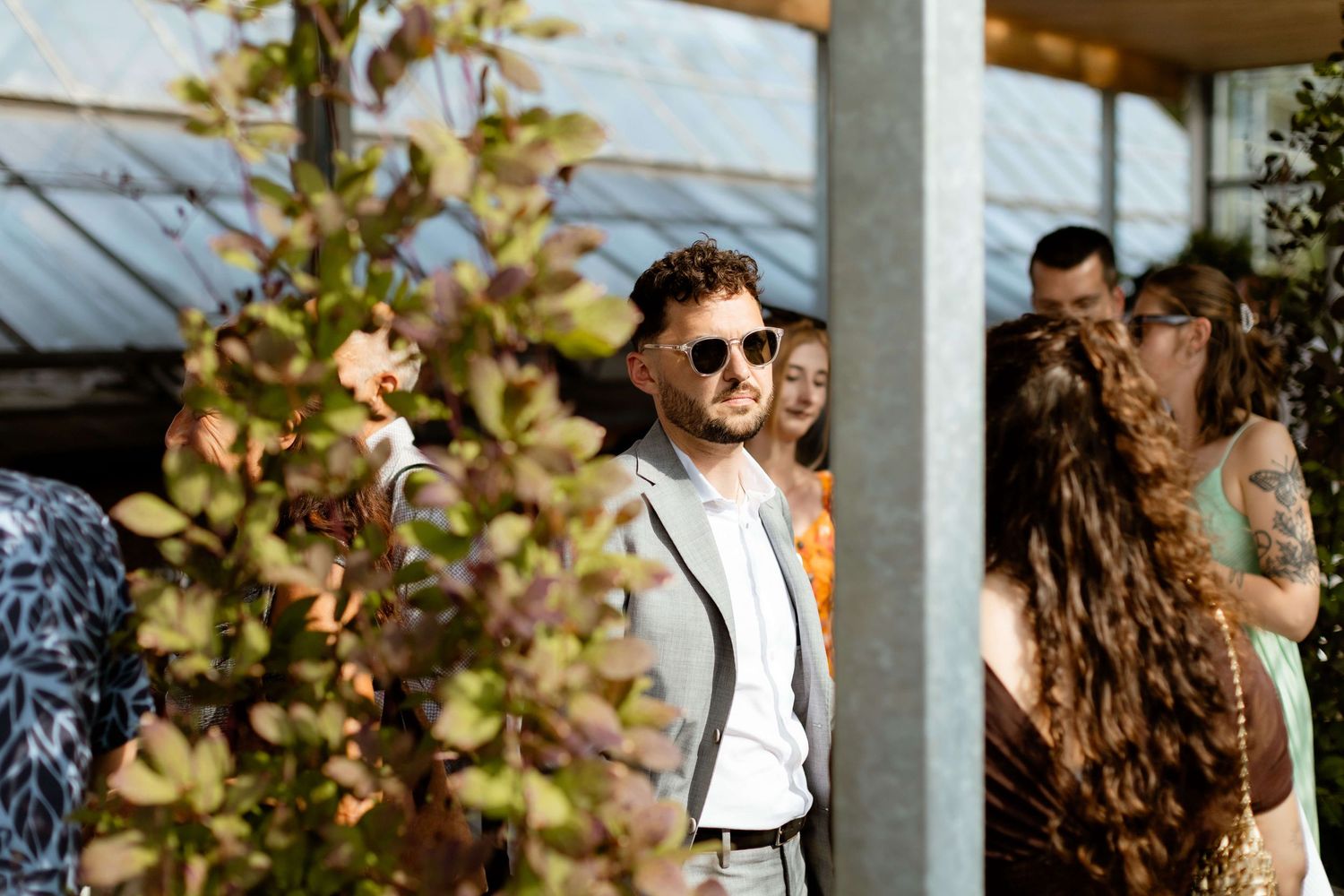 Stylish wedding guest in sunglasses standing among crowd outdoors.