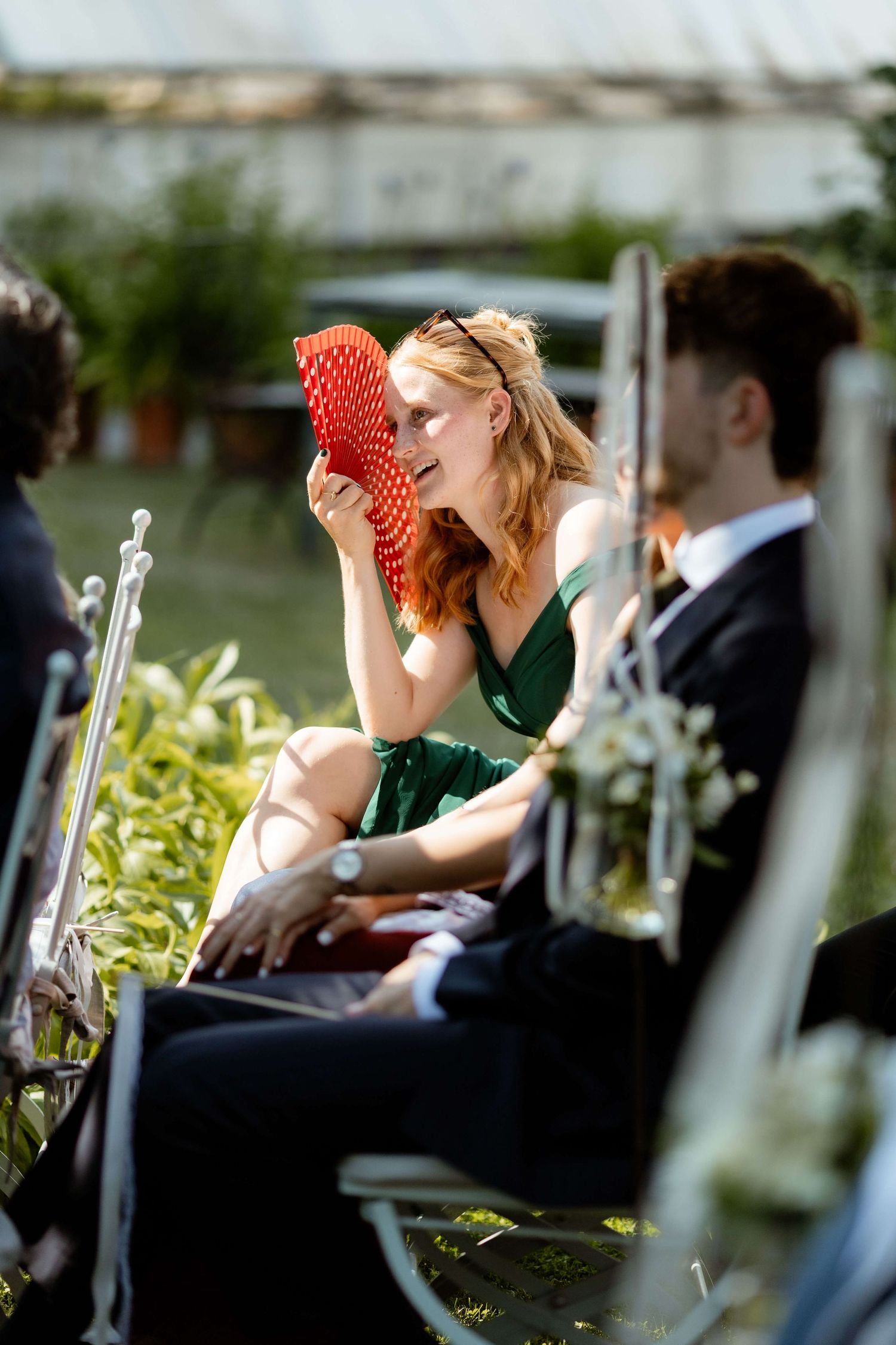 Smiling wedding guest in green dress fanning herself with red fan at sunny outdoor ceremony.