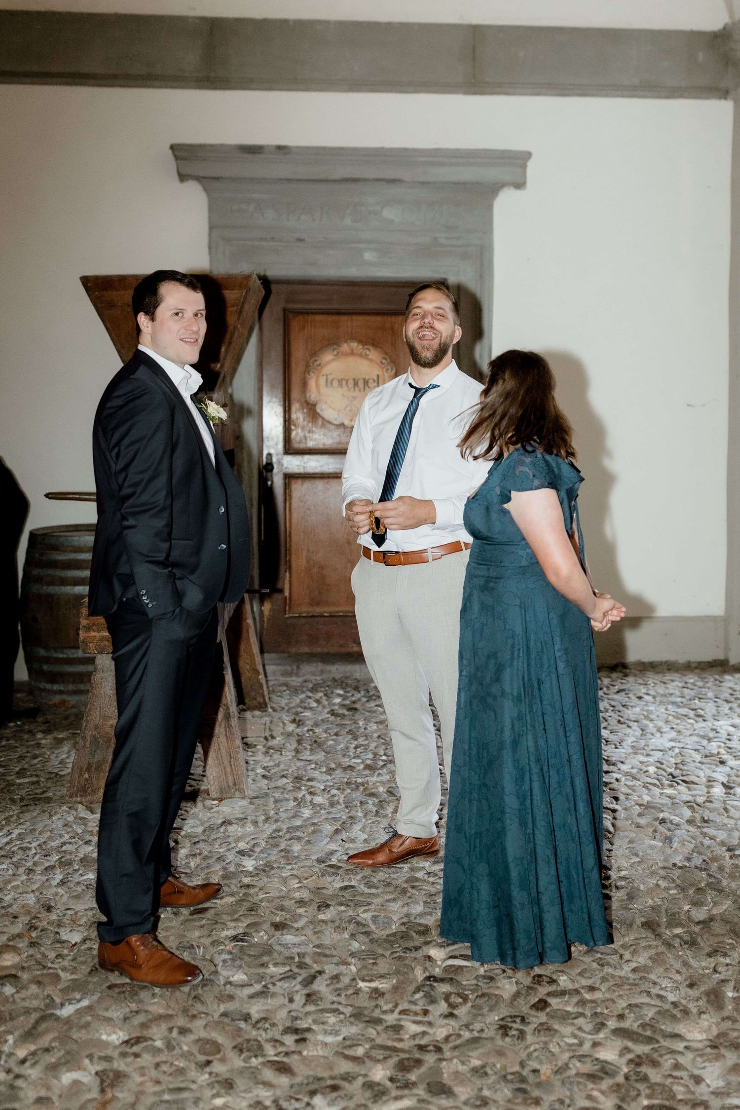 Three wedding guests chat and laugh together inside a rustic venue with wooden doors and wine barrels.