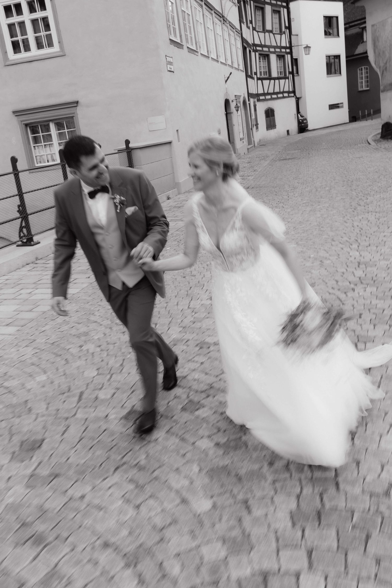 Black and white photo of happy bride and groom running hand-in-hand on cobblestone street.