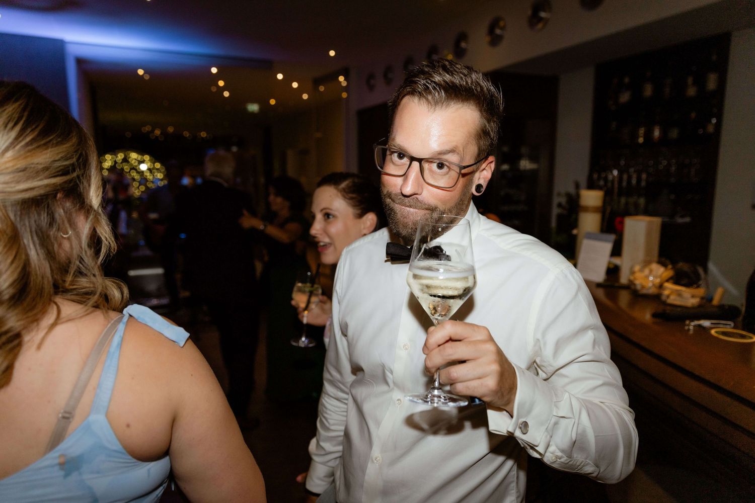 A bearded man wearing glasses raises a glass of white wine at an elegant indoor wedding reception.