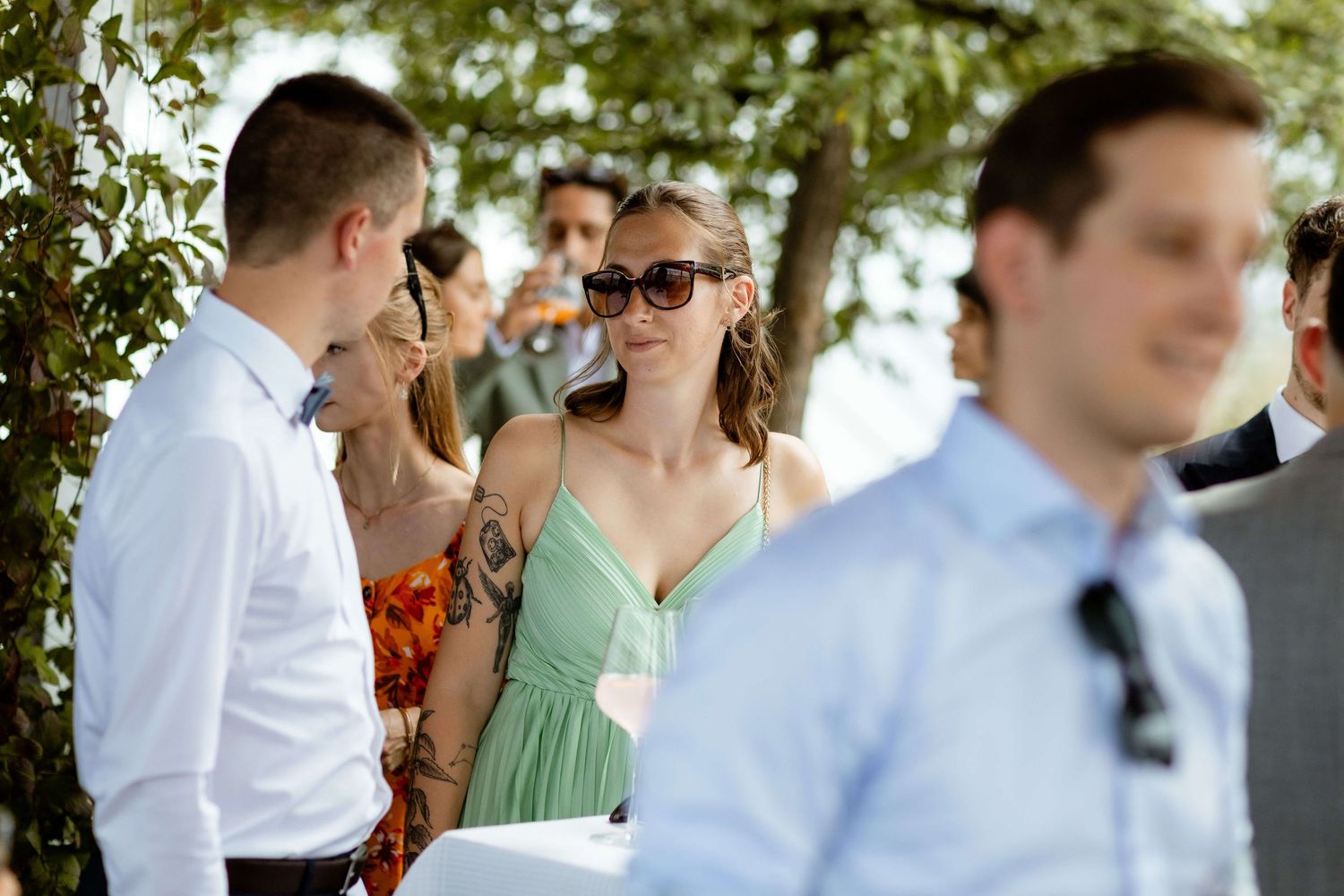 Wedding guests mingle outdoors, woman in green dress wearing sunglasses smiles.
