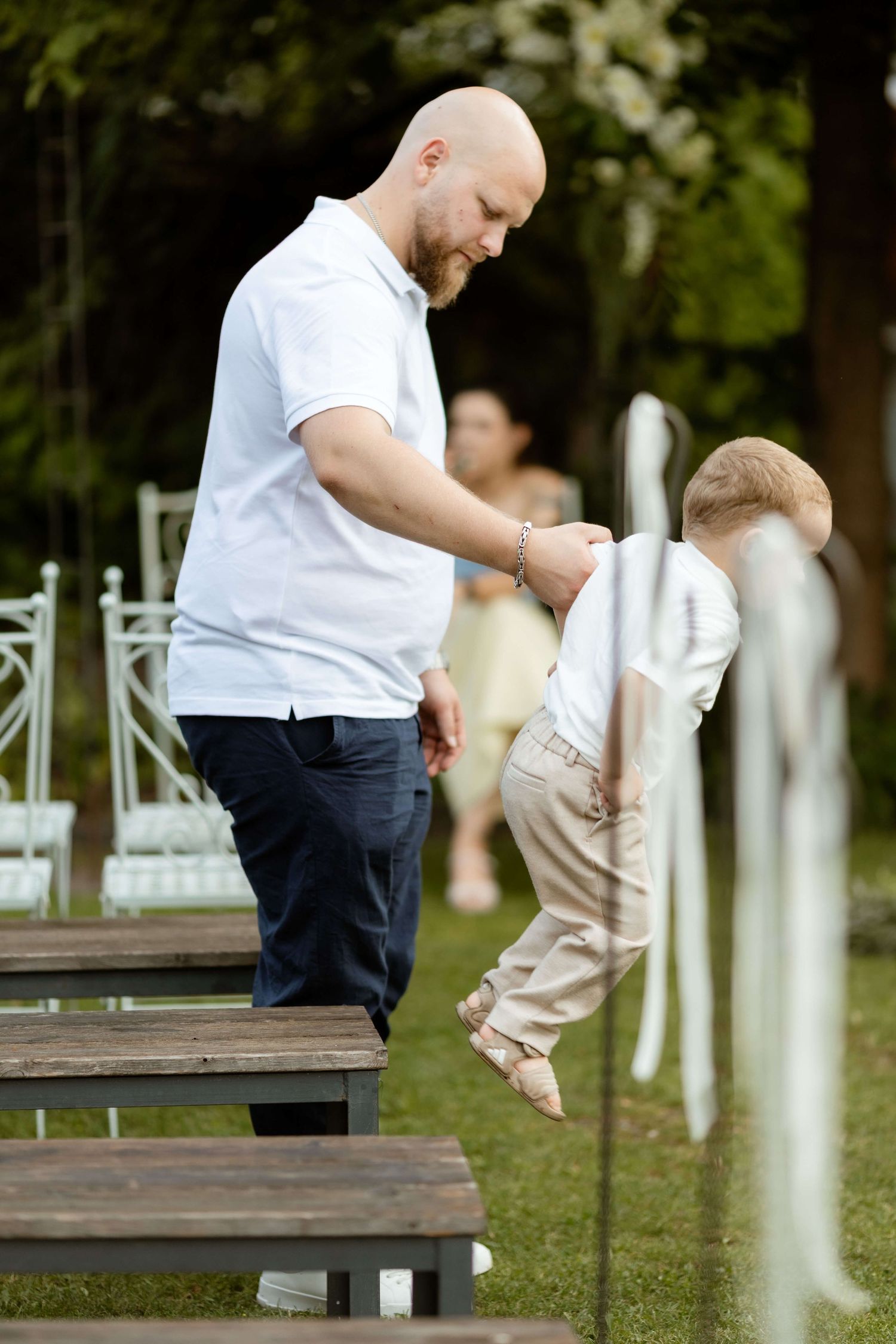 A bald man in a white polo shirt helps a small toddler balance on wooden benches at an outdoor wedding ceremony.
