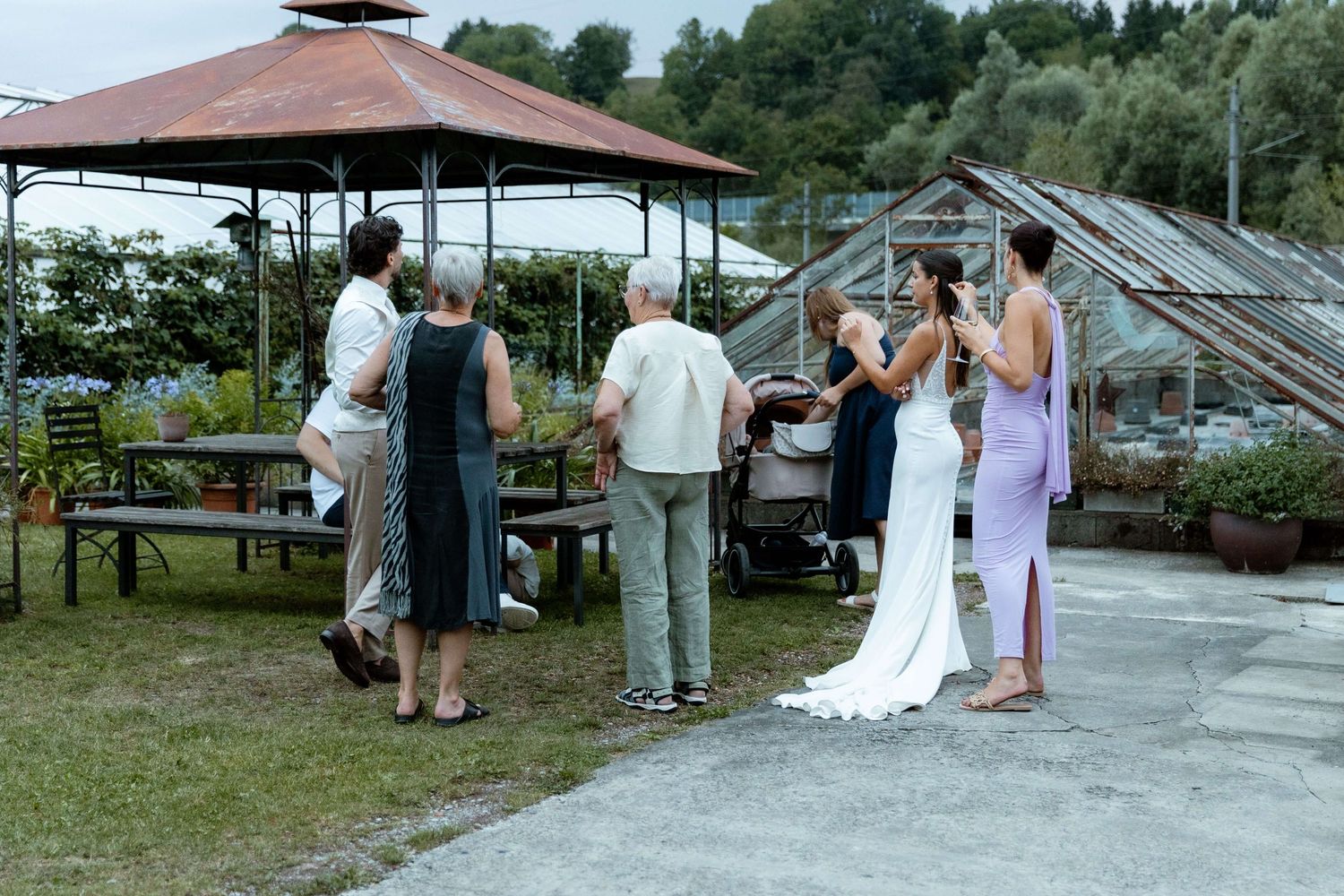 Wedding guests mingling with bride in white gown near rustic gazebo and greenhouse venue.