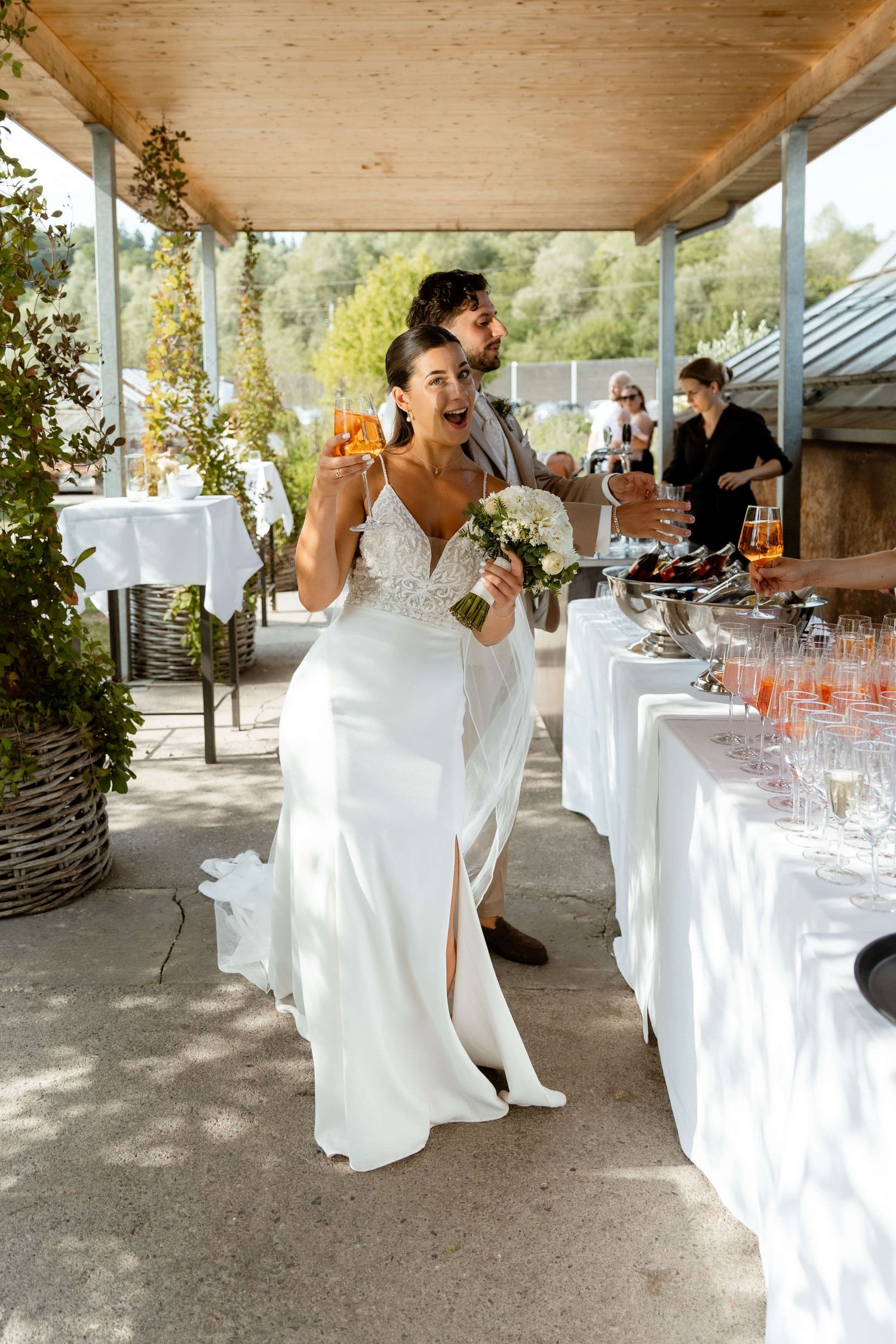 Bride in white gown holding bouquet and champagne at reception.