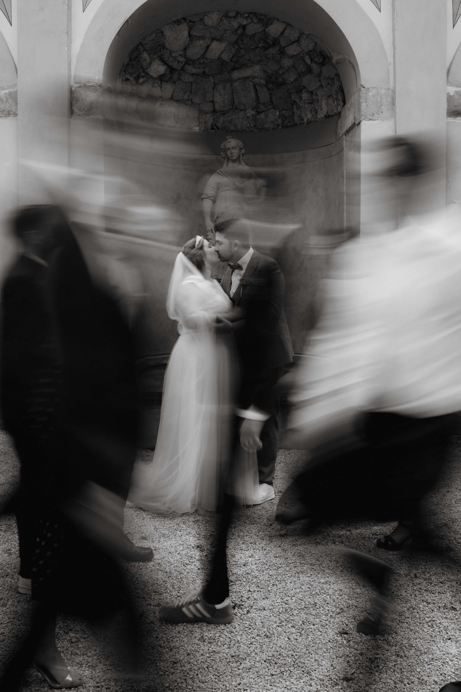 Artistic black and white blurred motion photo of bride and groom kissing amid moving wedding guests.