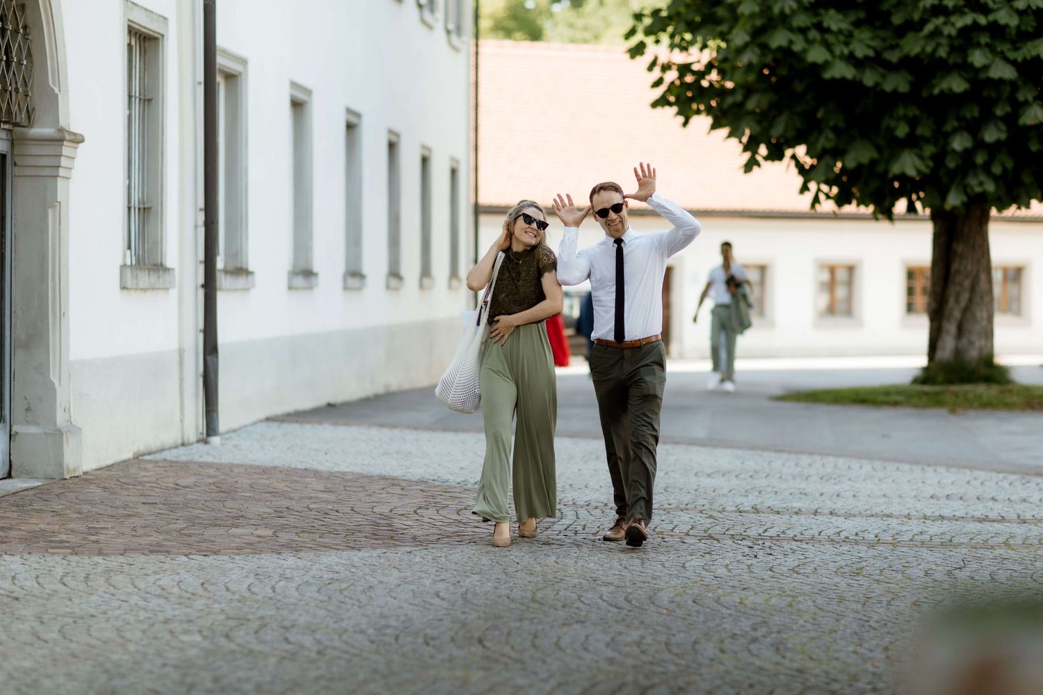 Couple walking hand in hand on cobblestone street, waving and smiling in a European town.