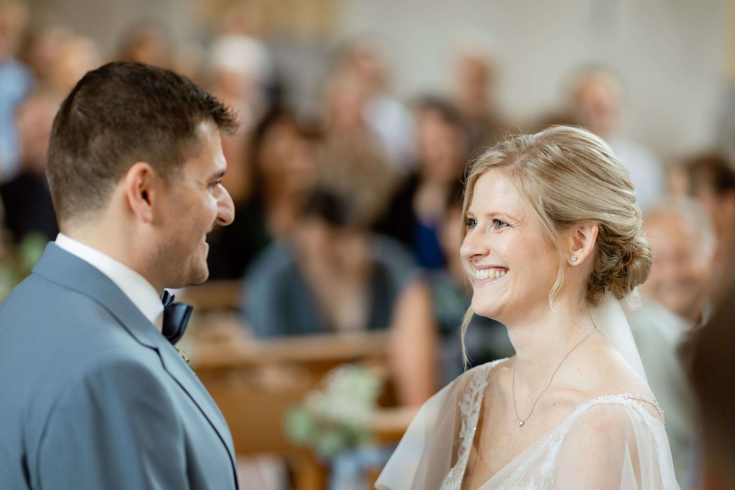 Smiling bride and groom exchanging vows at church wedding ceremony with guests behind.