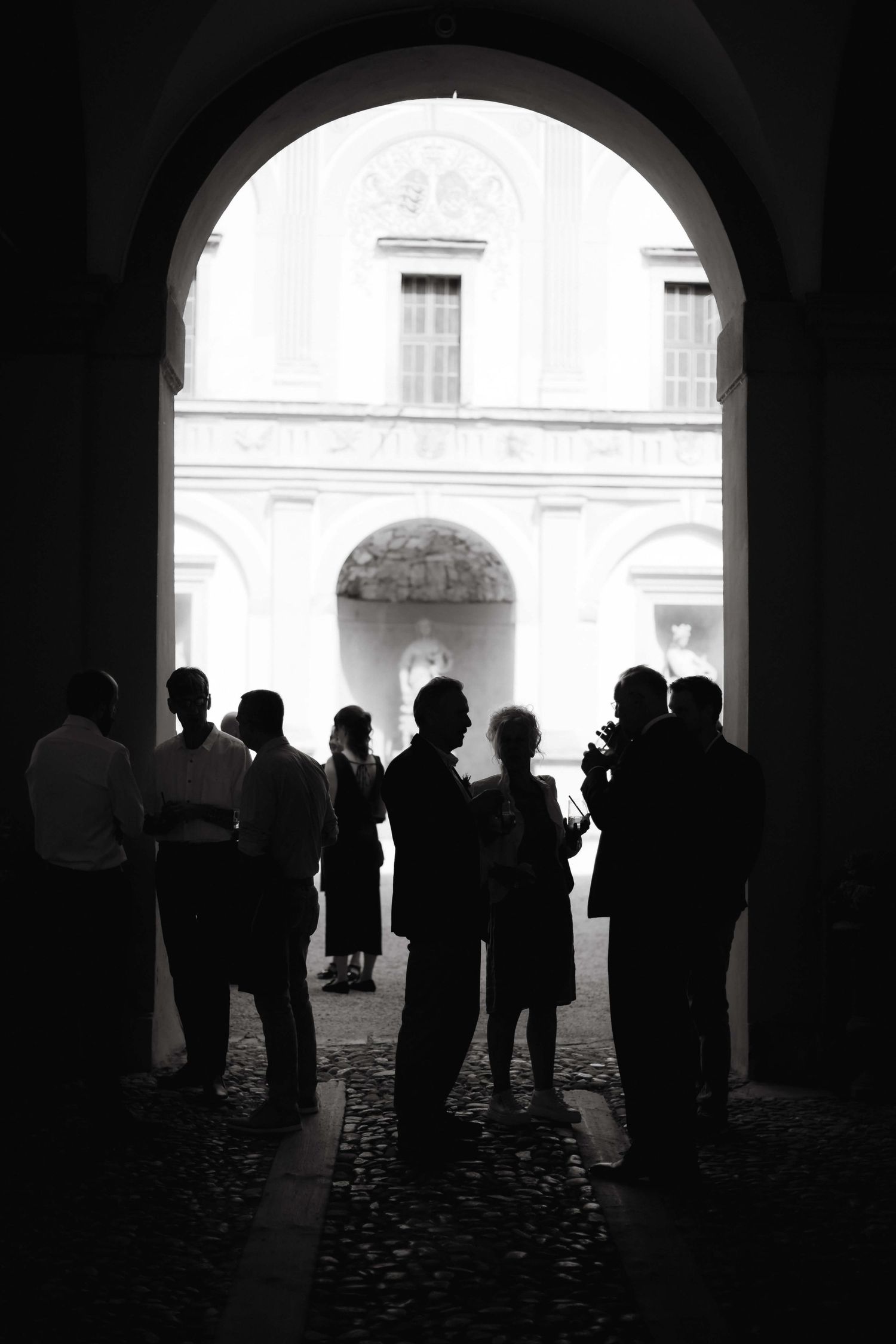 Black and white silhouette of wedding guests gathered under an arched entrance to a sunlit courtyard.