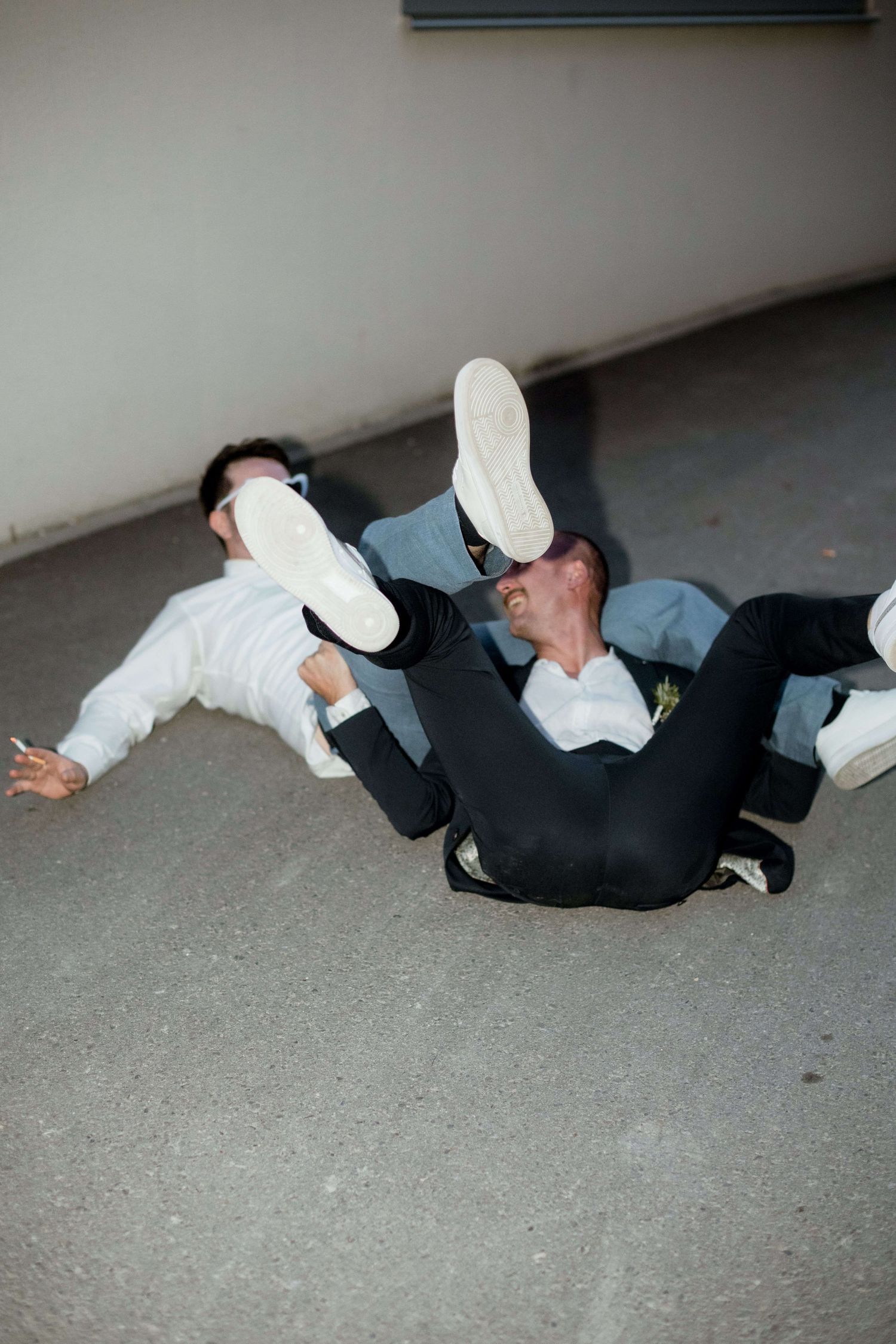 Two men in formal wear playfully wrestling on the ground outdoors at a wedding.