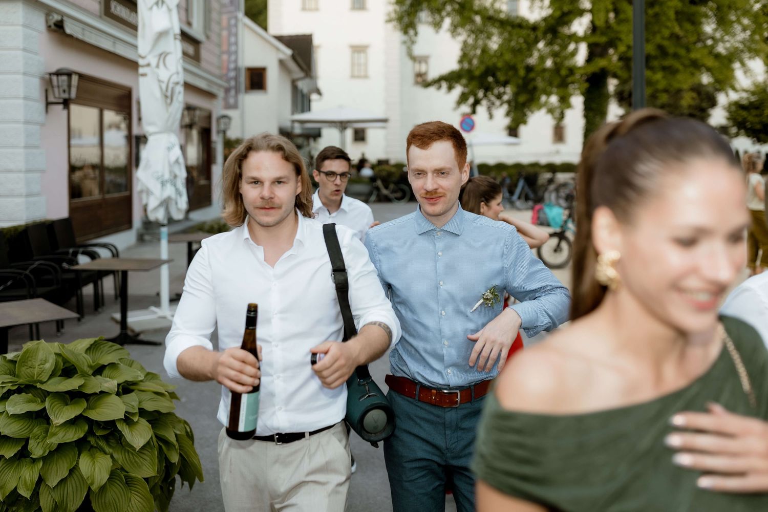 Two men carrying beer bottles walk along a sunny outdoor street during a wedding celebration.