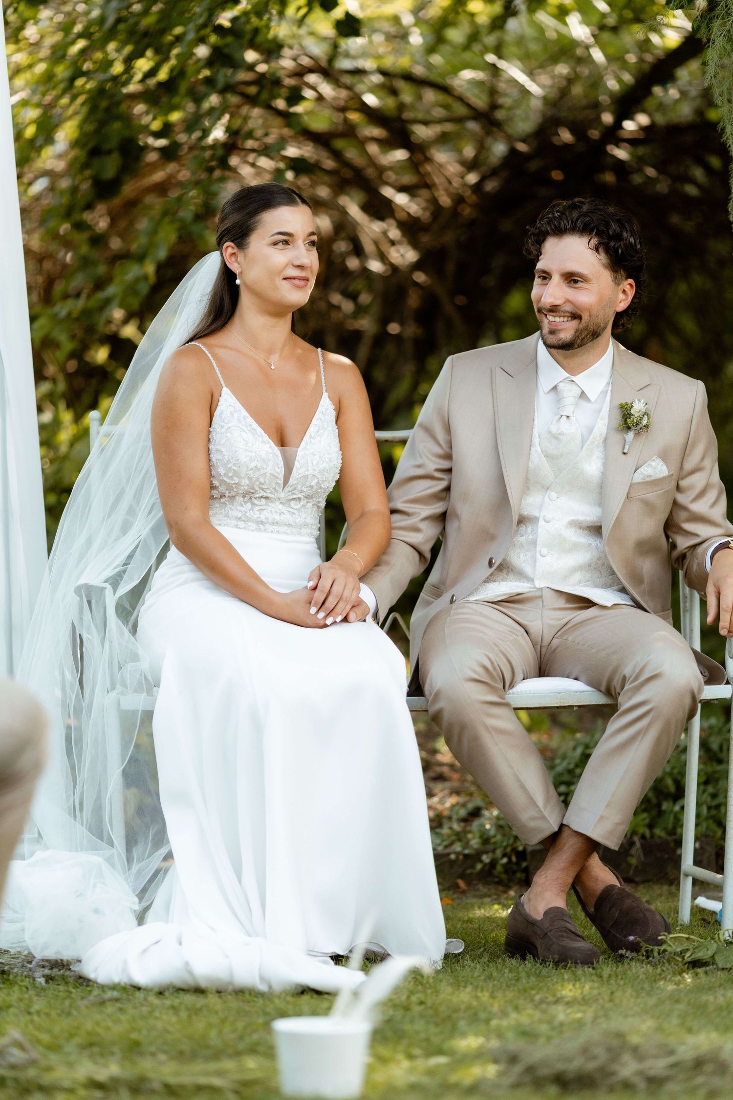 Smiling bride in white veil and groom in tan suit seated at outdoor garden wedding.