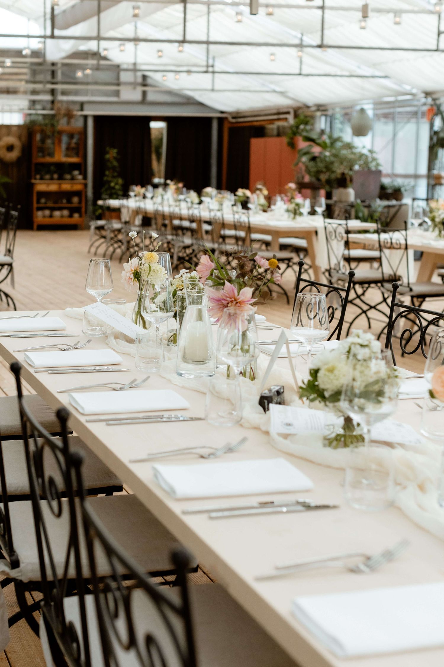 Long wedding reception table with pastel floral centerpieces in a greenhouse venue.