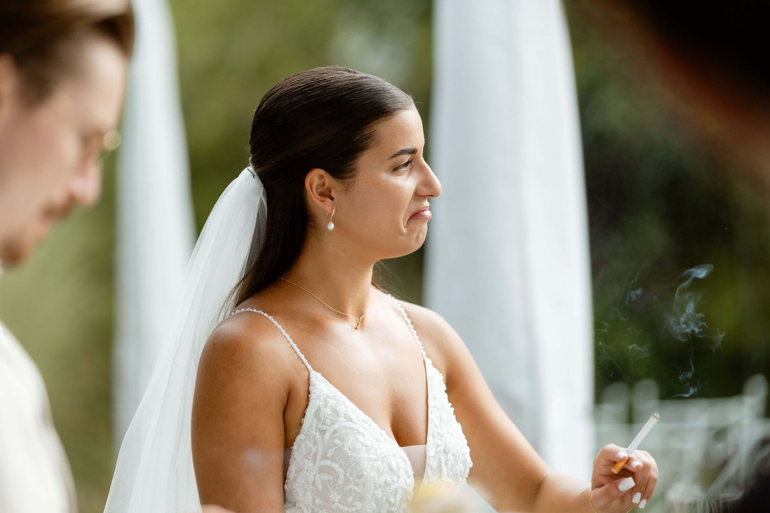 Elegant bride in white lace dress and veil holding a cigarette at outdoor wedding.