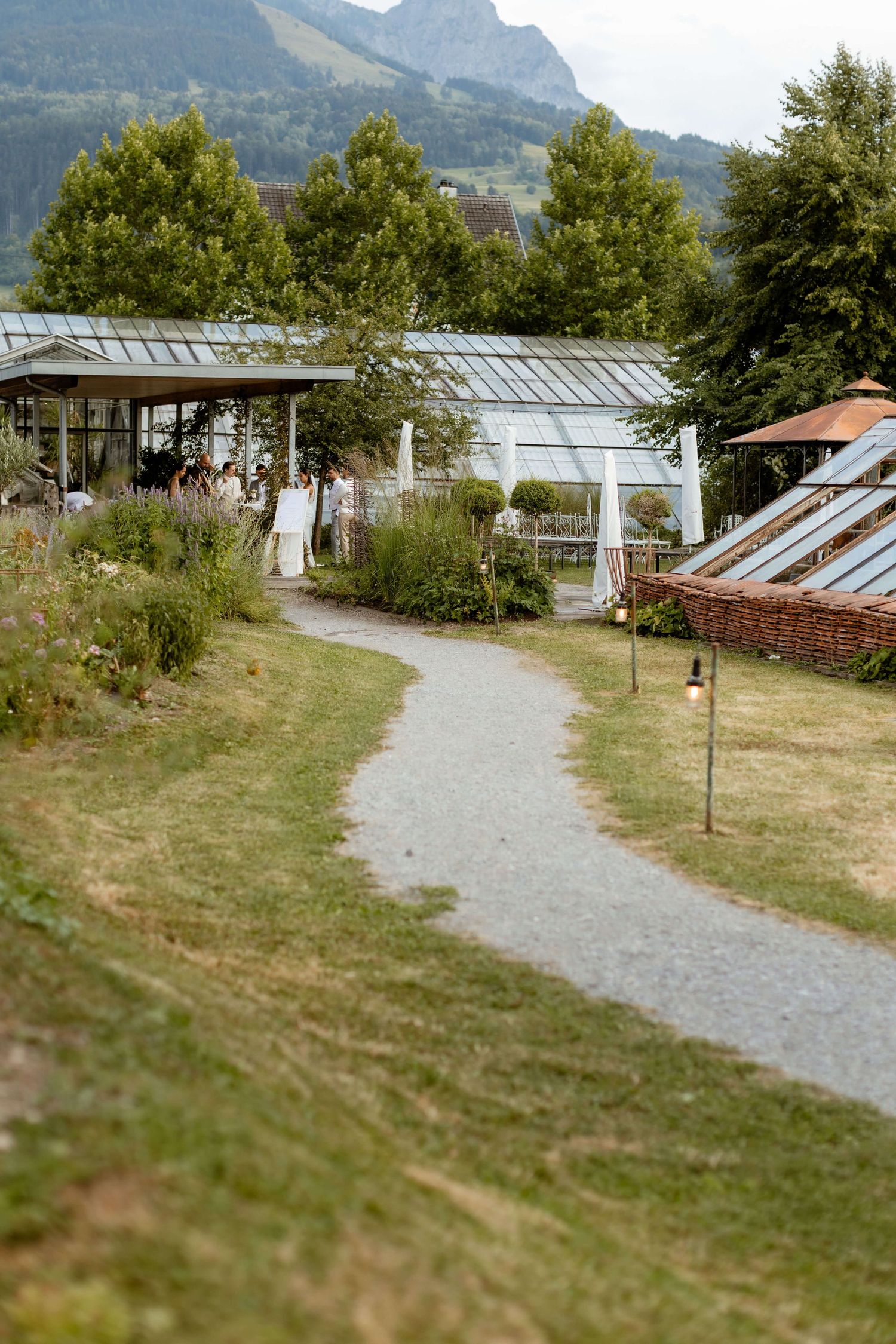 Winding gravel path leading to greenhouse wedding venue surrounded by lush gardens and mountains.