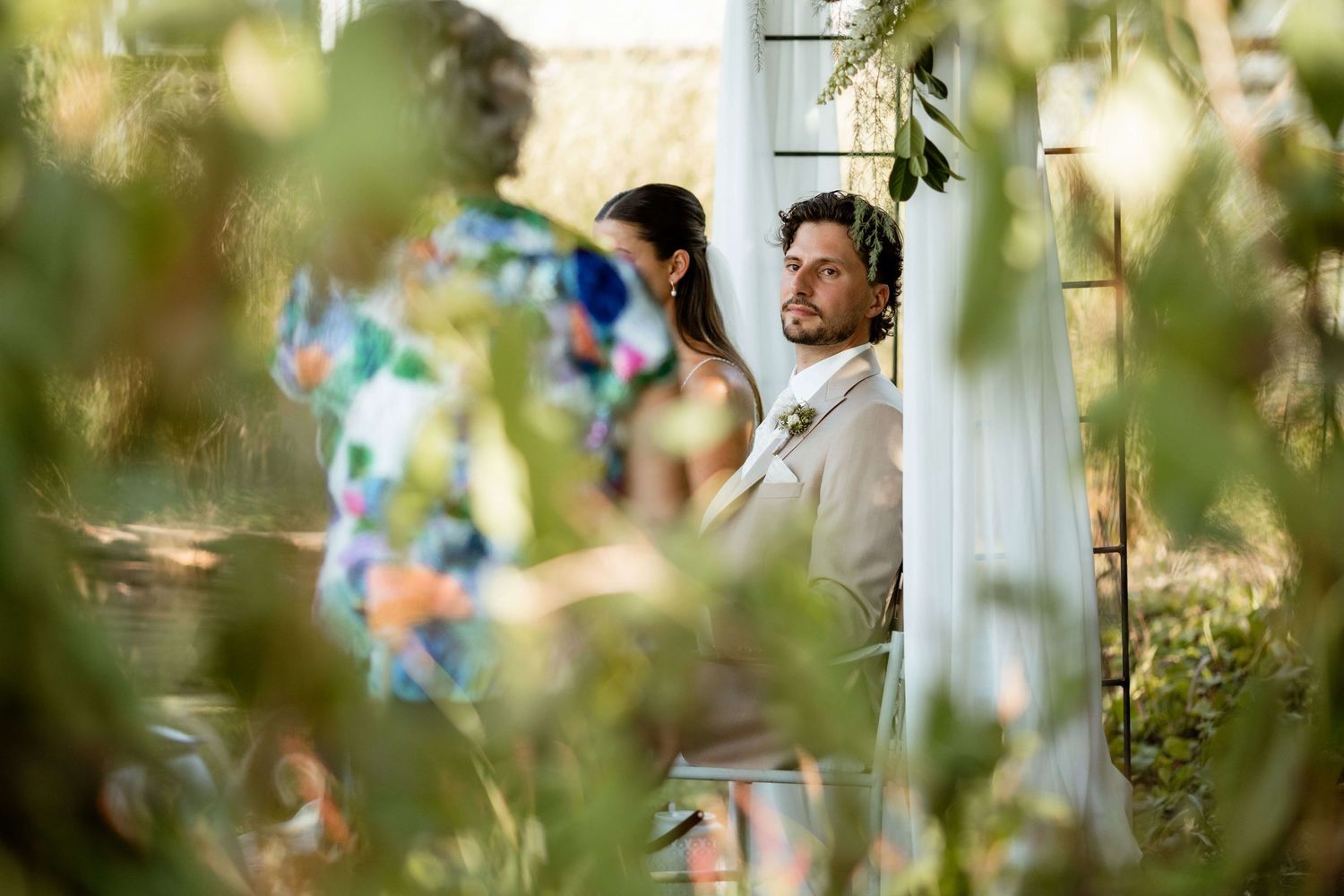 Groom in beige suit peeks around white pillar surrounded by lush greenery at wedding.