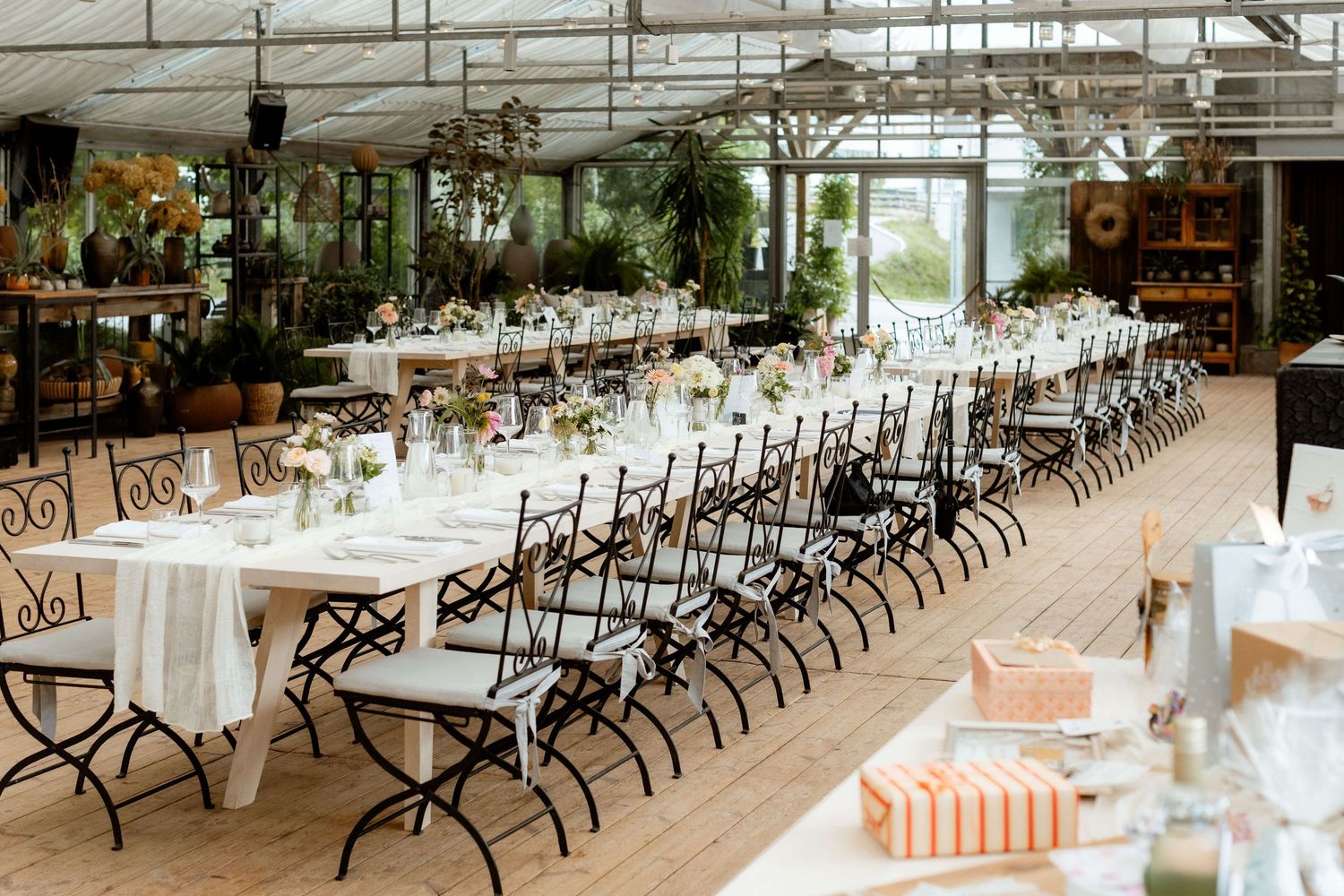 Elegant greenhouse wedding reception with long white tables, floral centerpieces, and wrought iron chairs on wooden floors.