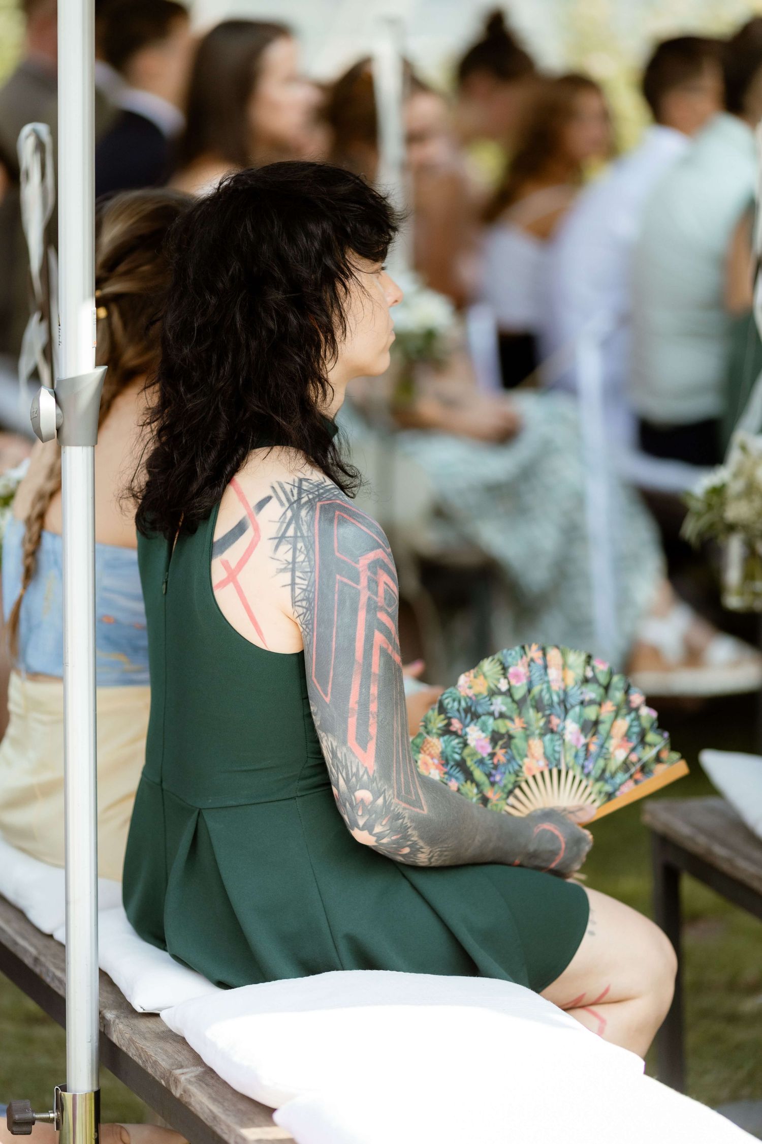 Wedding guest in green dress with tattoo sleeve holds decorative fan at outdoor ceremony.