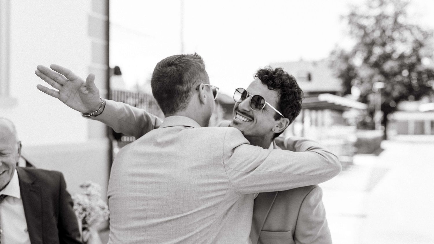 Two men sharing a warm embrace and greeting at an outdoor wedding celebration in black and white.