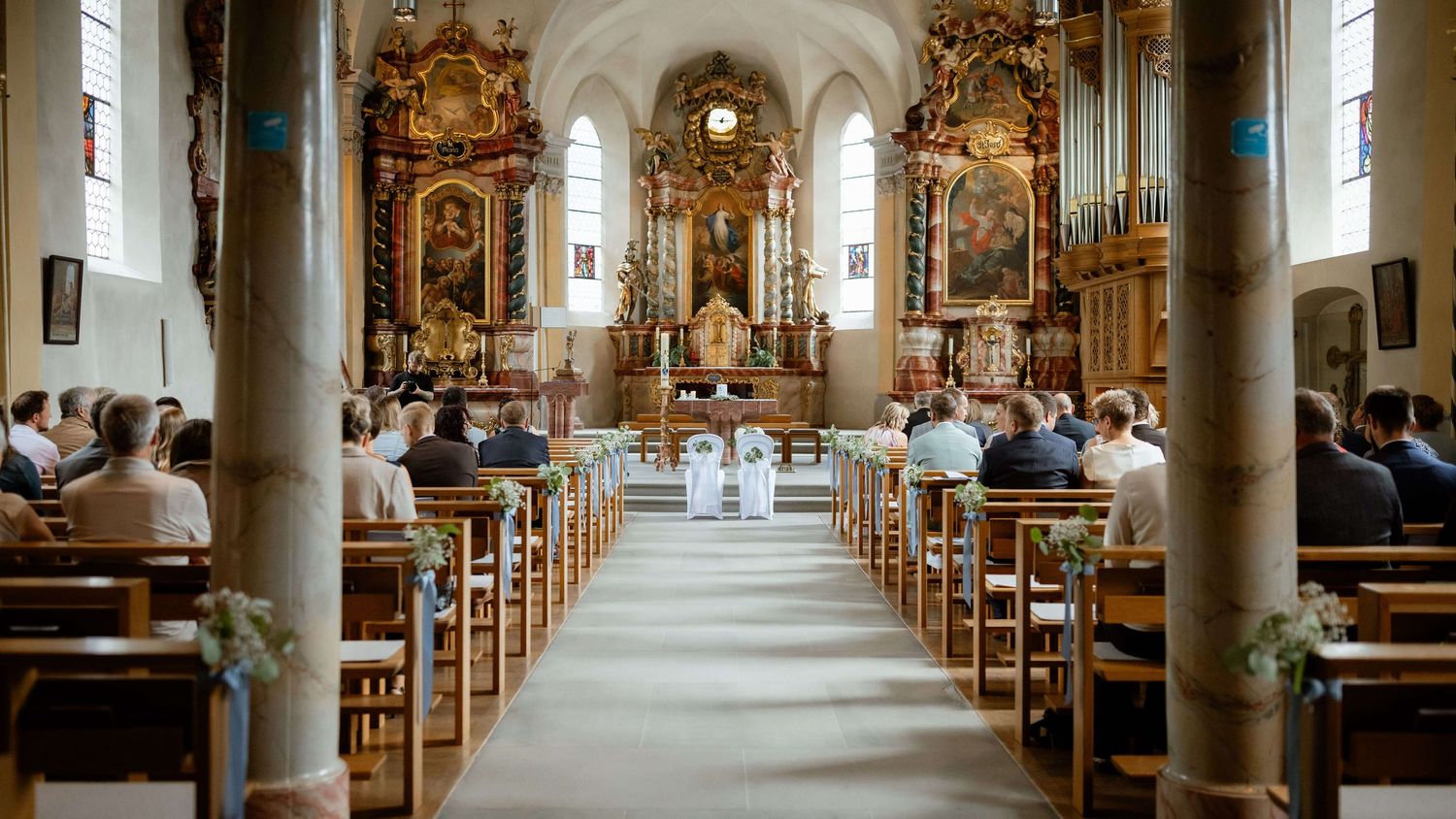 Wide view of ornate church interior filled with wedding guests seated in wooden pews.