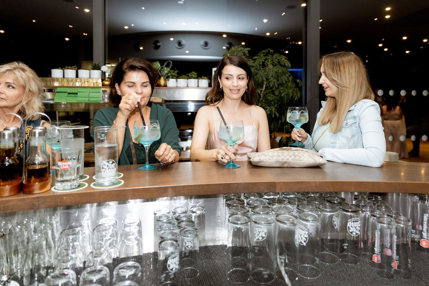 Four women sit at a bar counter enjoying cocktails with gin glasses and drinks at an event.