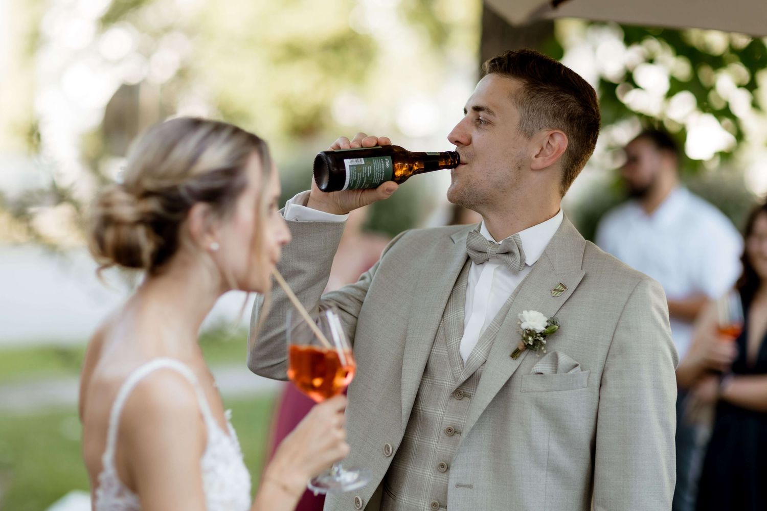 Groom in beige suit drinks beer bottle while bride holds rosé at wedding reception outdoors.