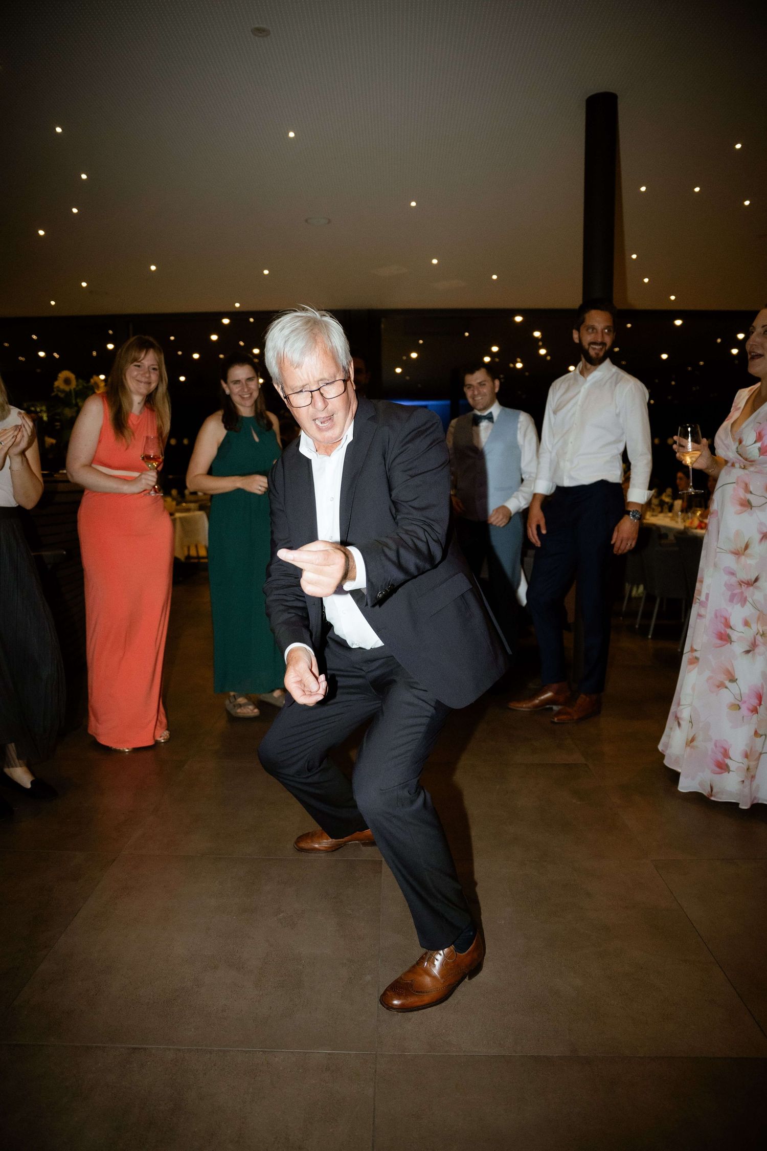 An older man in a dark suit shows off energetic dance moves on a wedding reception dance floor.