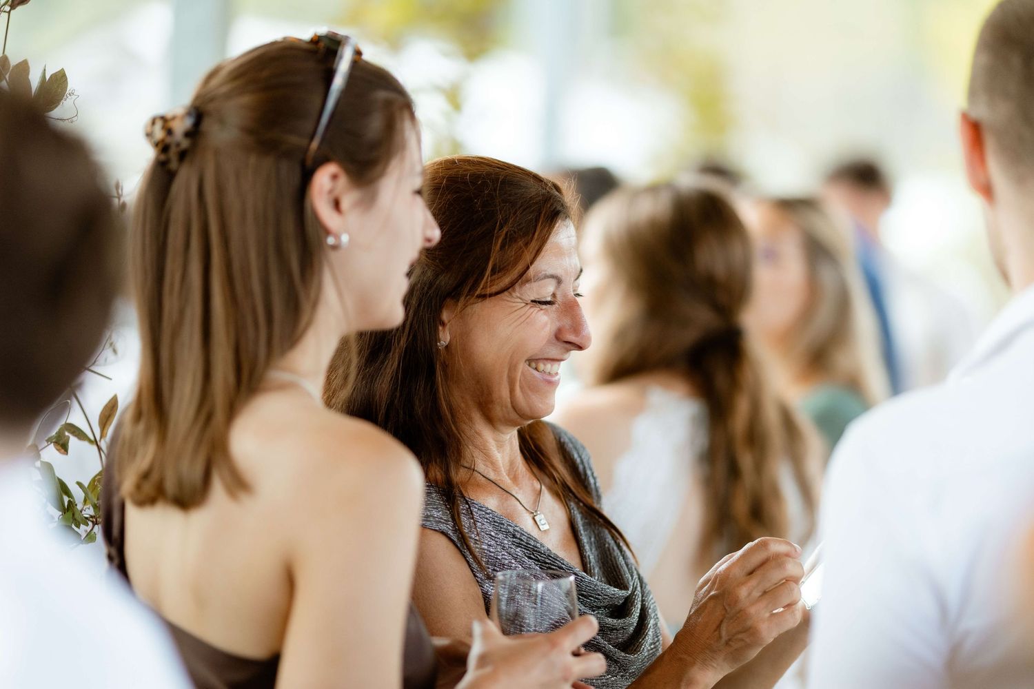 Two women share a joyful laugh together at a social gathering, one wearing a silver dress and the other in brown.