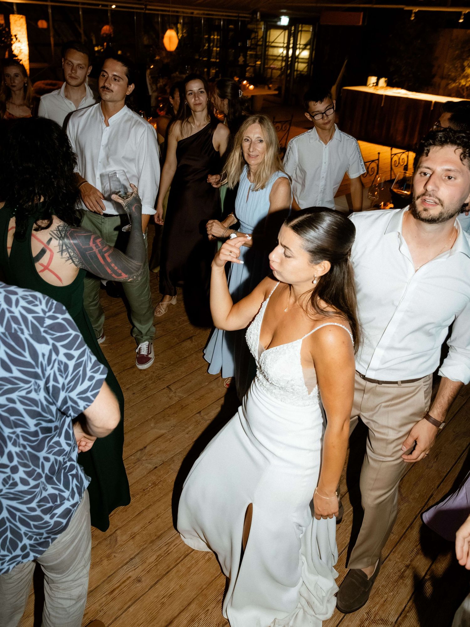 Bride in white spaghetti-strap gown dancing with guests at vibrant wedding reception.