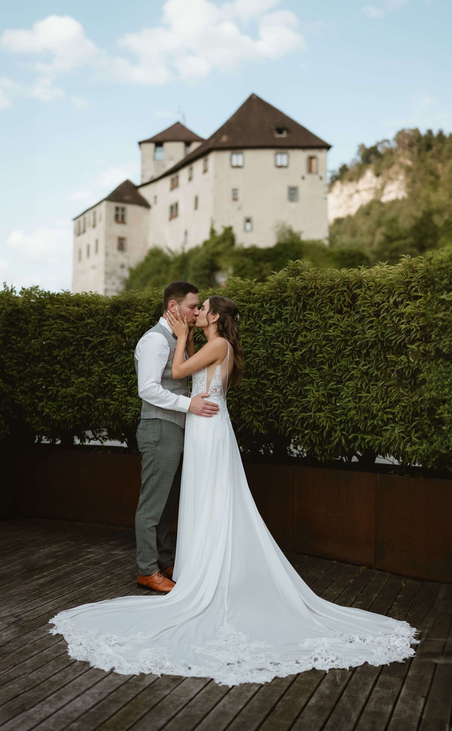 Bride and groom kissing on terrace with historic castle and hedges behind them.
