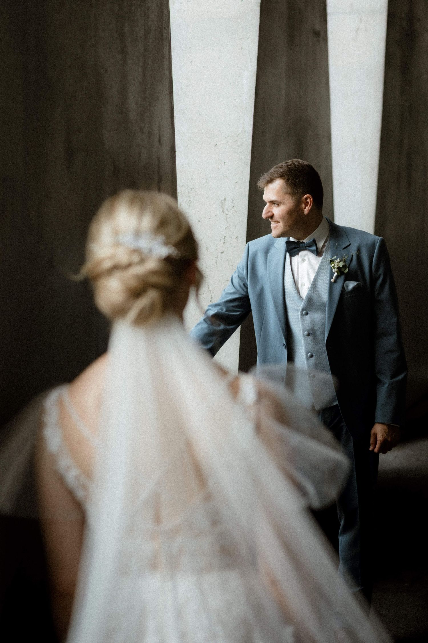 Groom in blue suit gazes at bride in lace wedding dress near large windows in soft light.