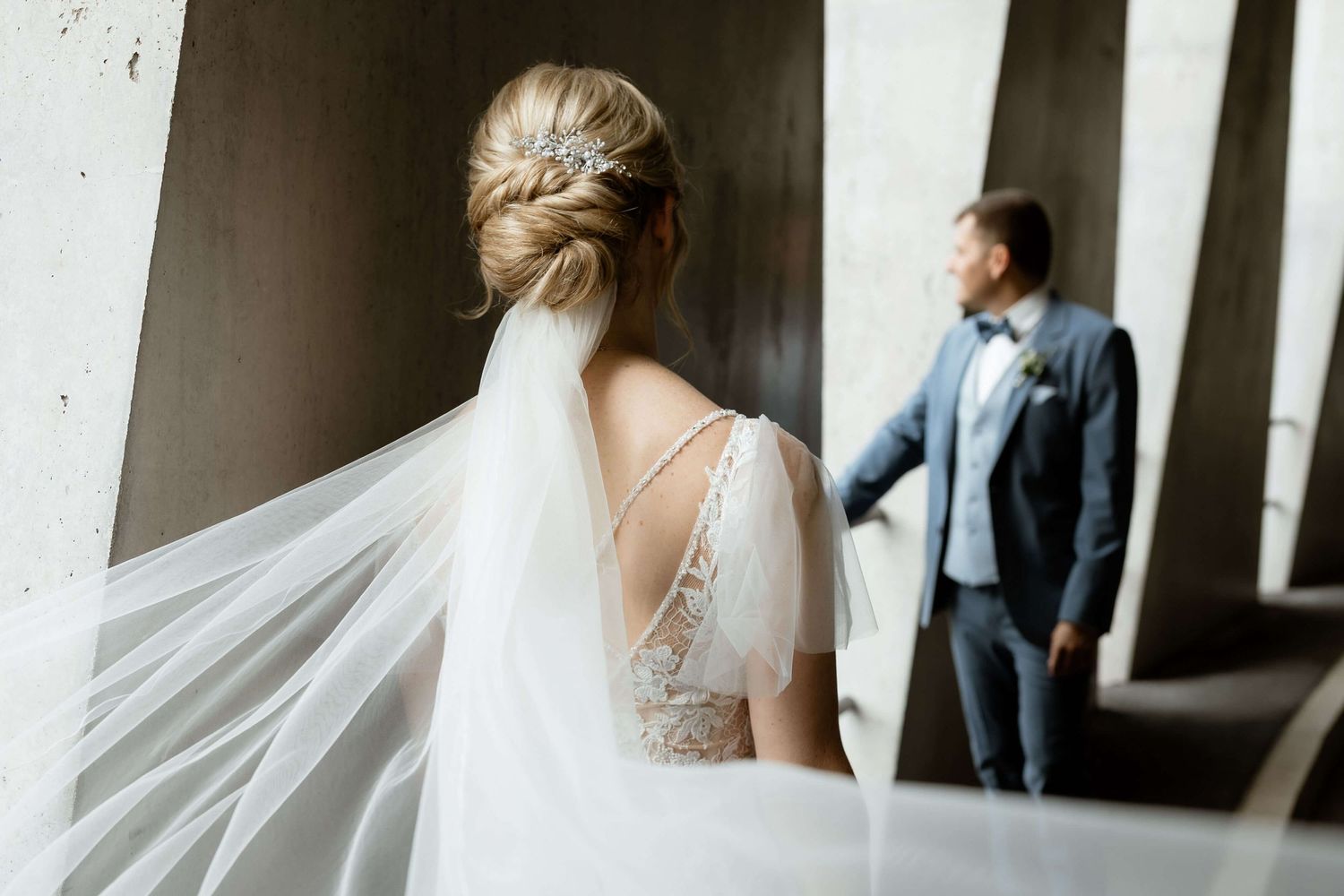 Bride in elegant lace back dress with long flowing veil walking toward groom in navy suit.