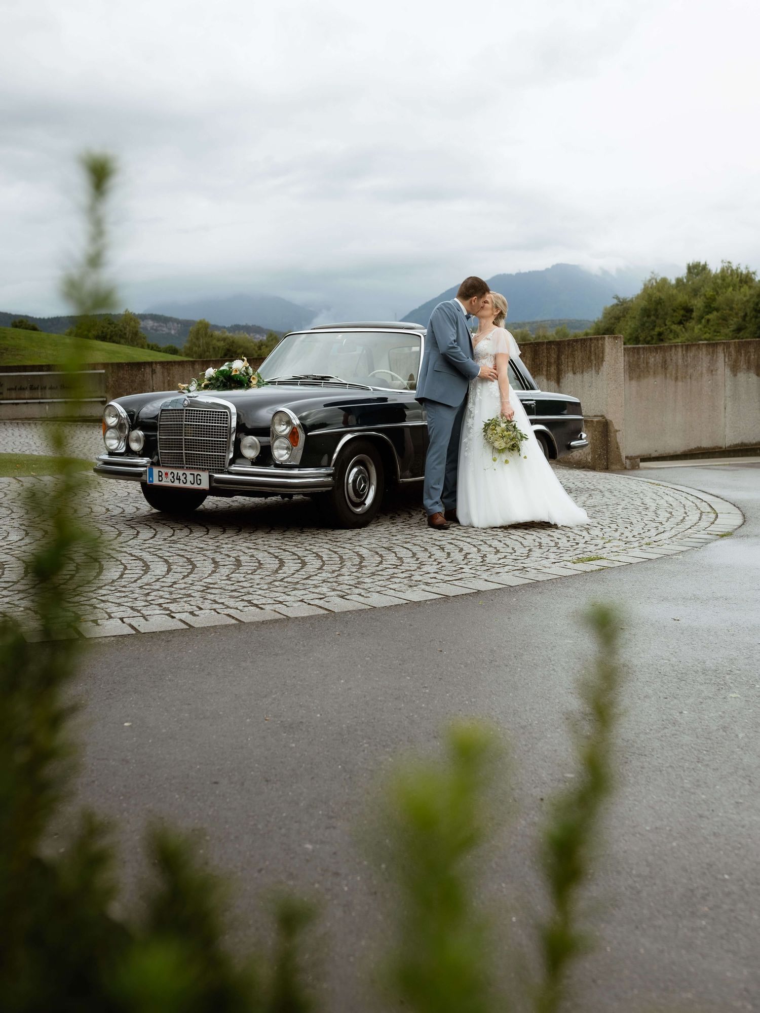 Bride and groom kiss beside a classic black Mercedes on cobblestone with mountain views.