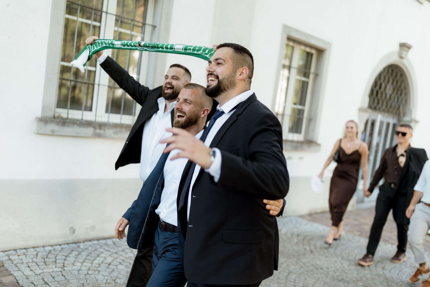 Groomsmen celebrating outdoors, one waving a green scarf, laughing and walking together after a wedding ceremony.