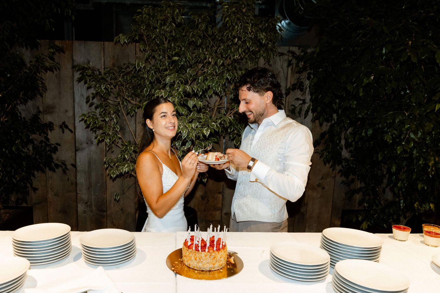 Happy couple sharing first bite of wedding cake at outdoor reception table at night.