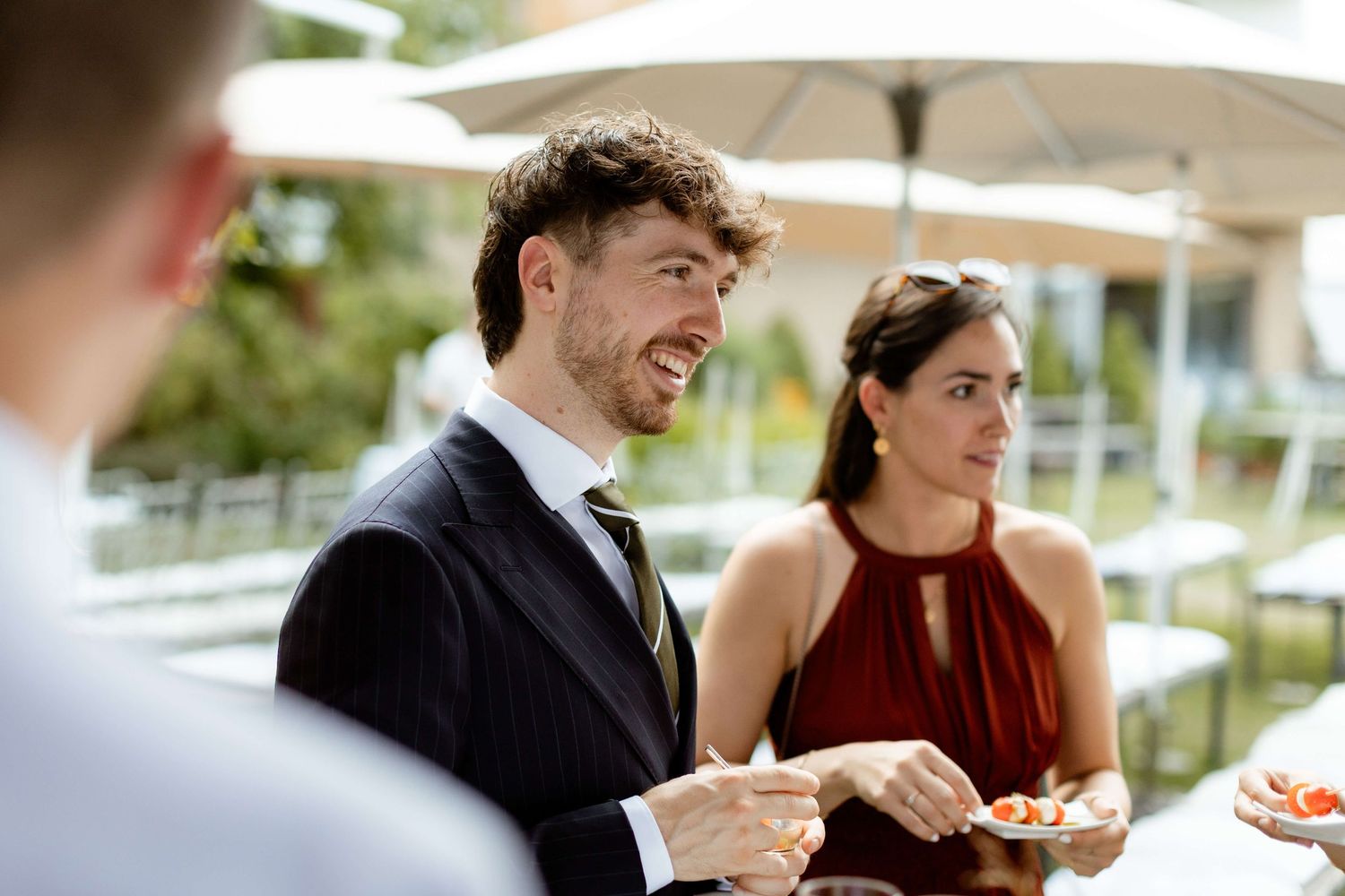 Smiling man in dark suit and woman in burgundy dress enjoy food at outdoor wedding.