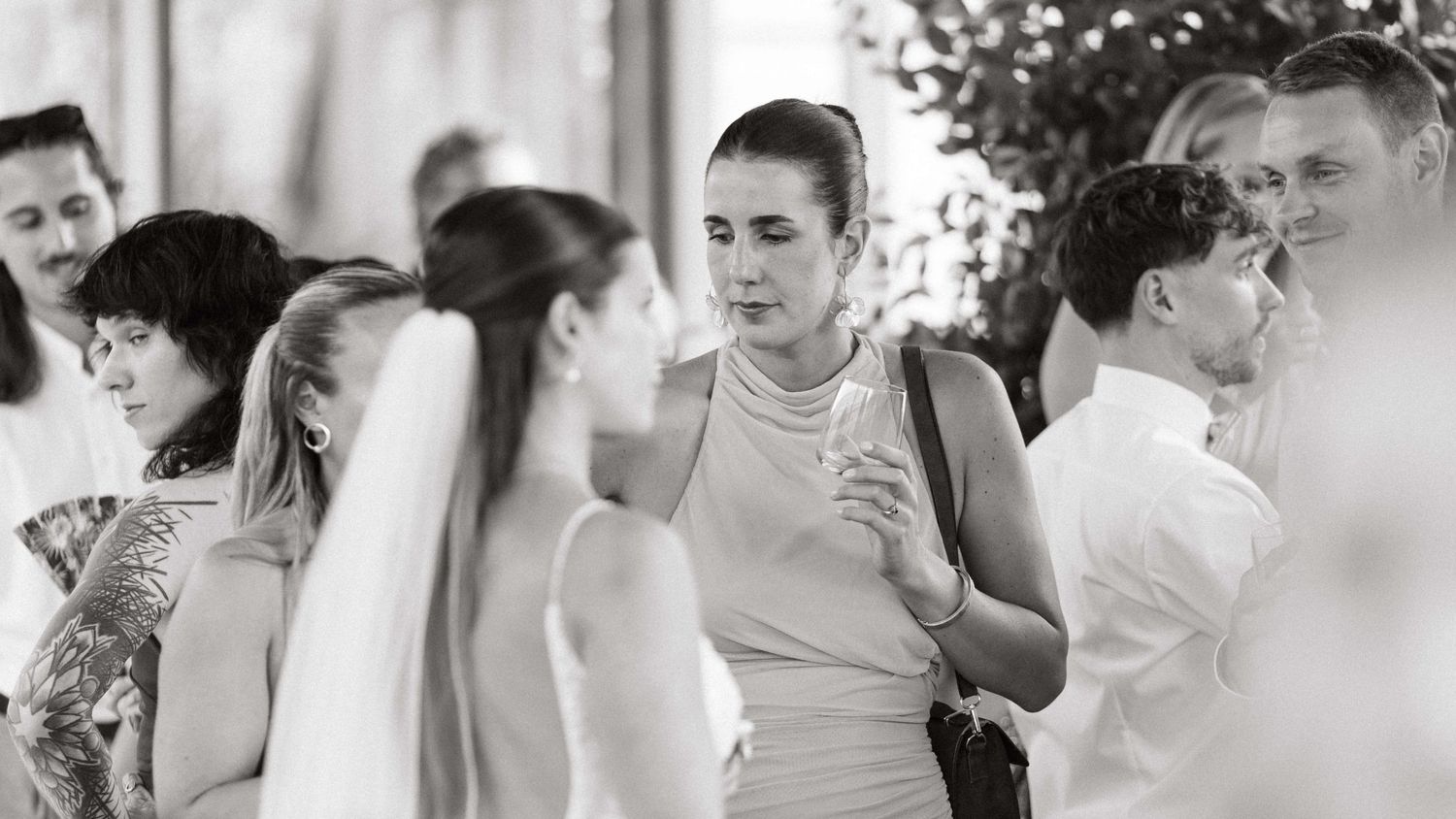 Black and white photo of wedding guests mingling, featuring a bride in a veil and a woman in a sleeveless dress.