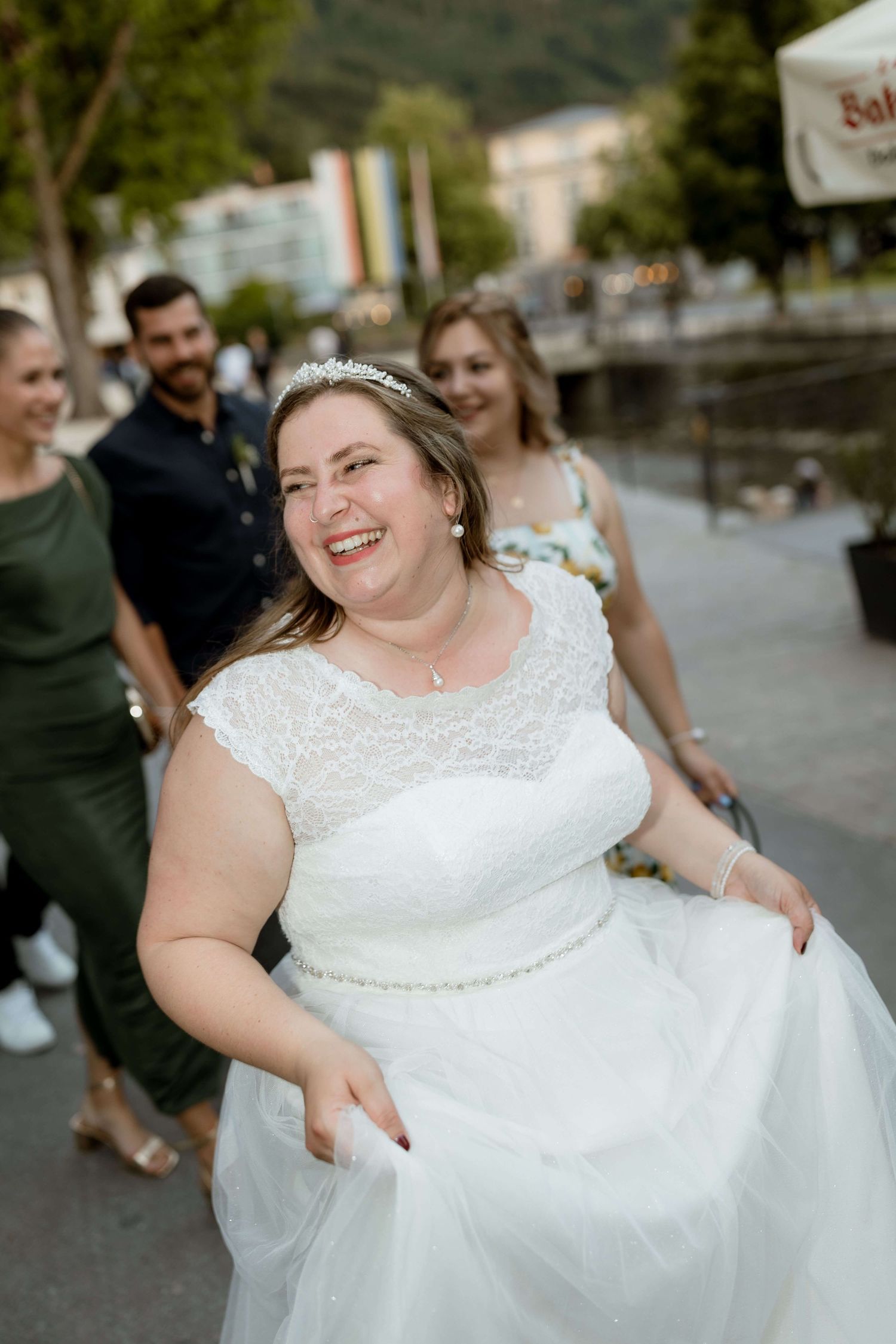 Radiant bride in a white lace dress and tiara smiles while walking outdoors with wedding guests behind her.
