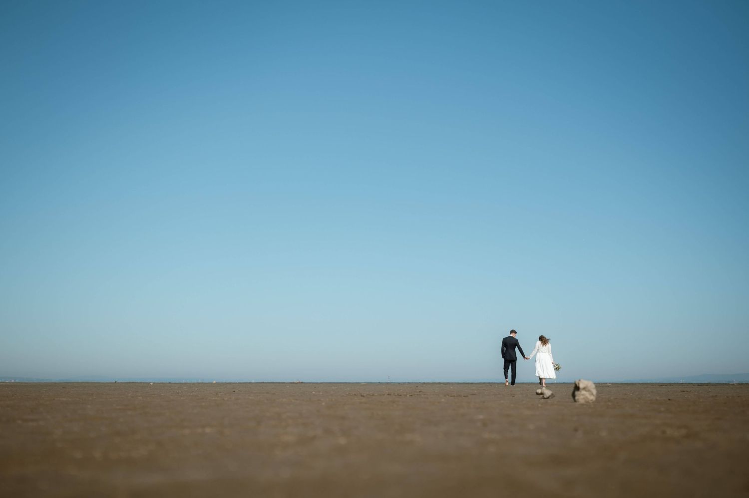 Bride and groom walking together on vast open sandy beach under clear blue sky.