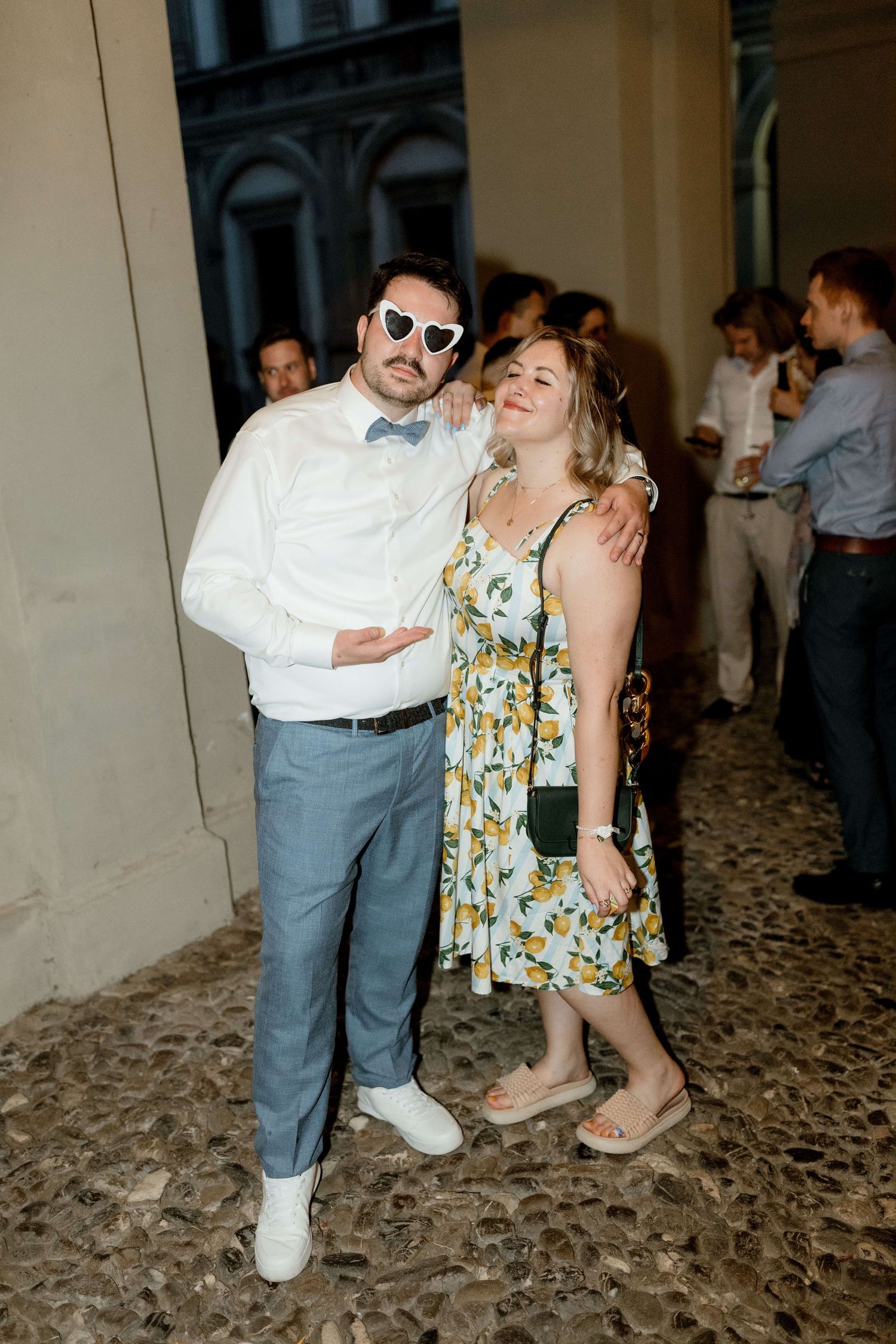 Man in heart sunglasses and bow tie posing with smiling woman in floral dress outdoors.