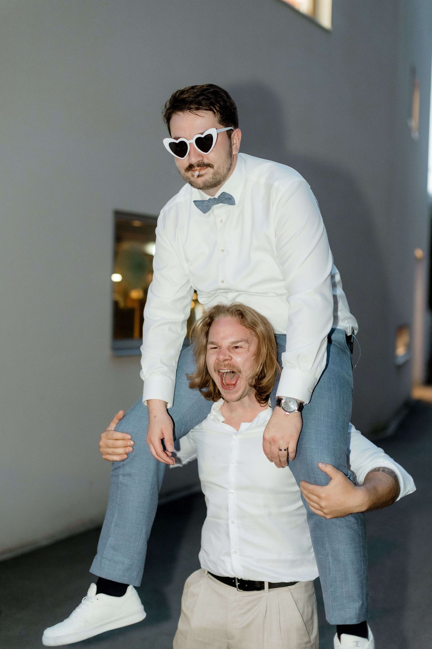 Man wearing heart sunglasses sitting on laughing long-haired mans shoulders indoors.
