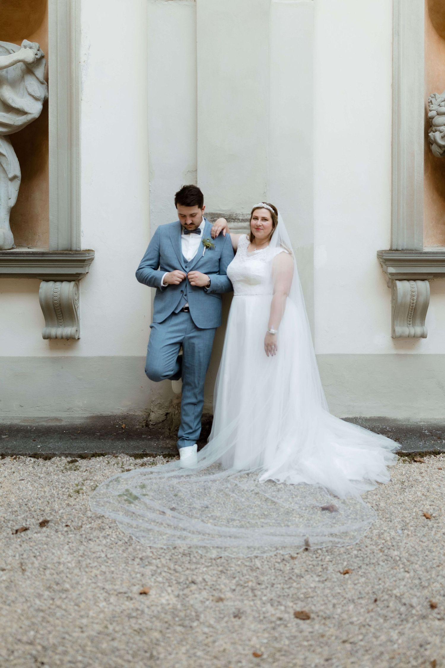 Bride in elegant white gown with long veil standing beside groom in blue suit at ornate venue.