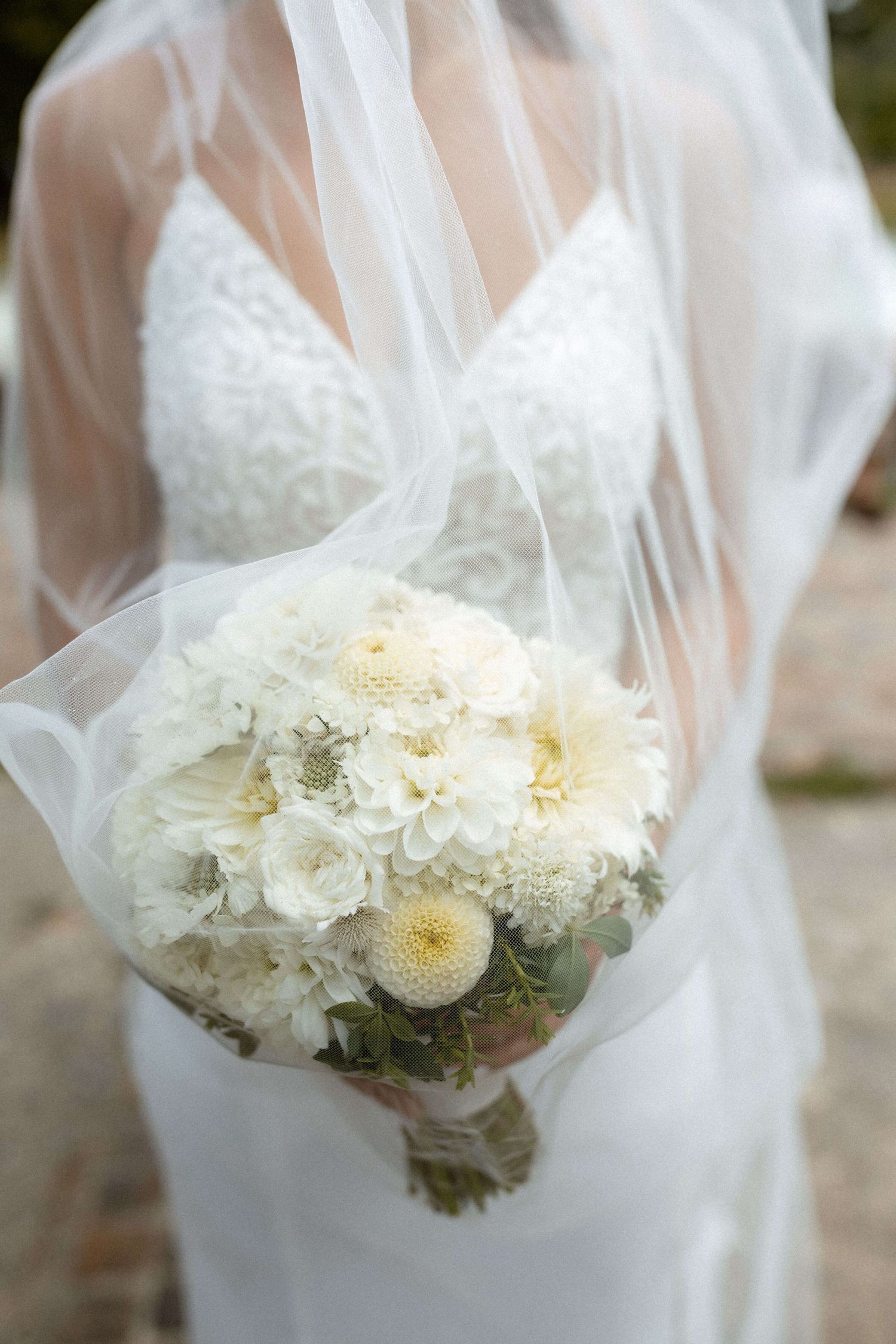 Bride in white lace gown holding a lush white dahlia bouquet draped with a flowing veil.