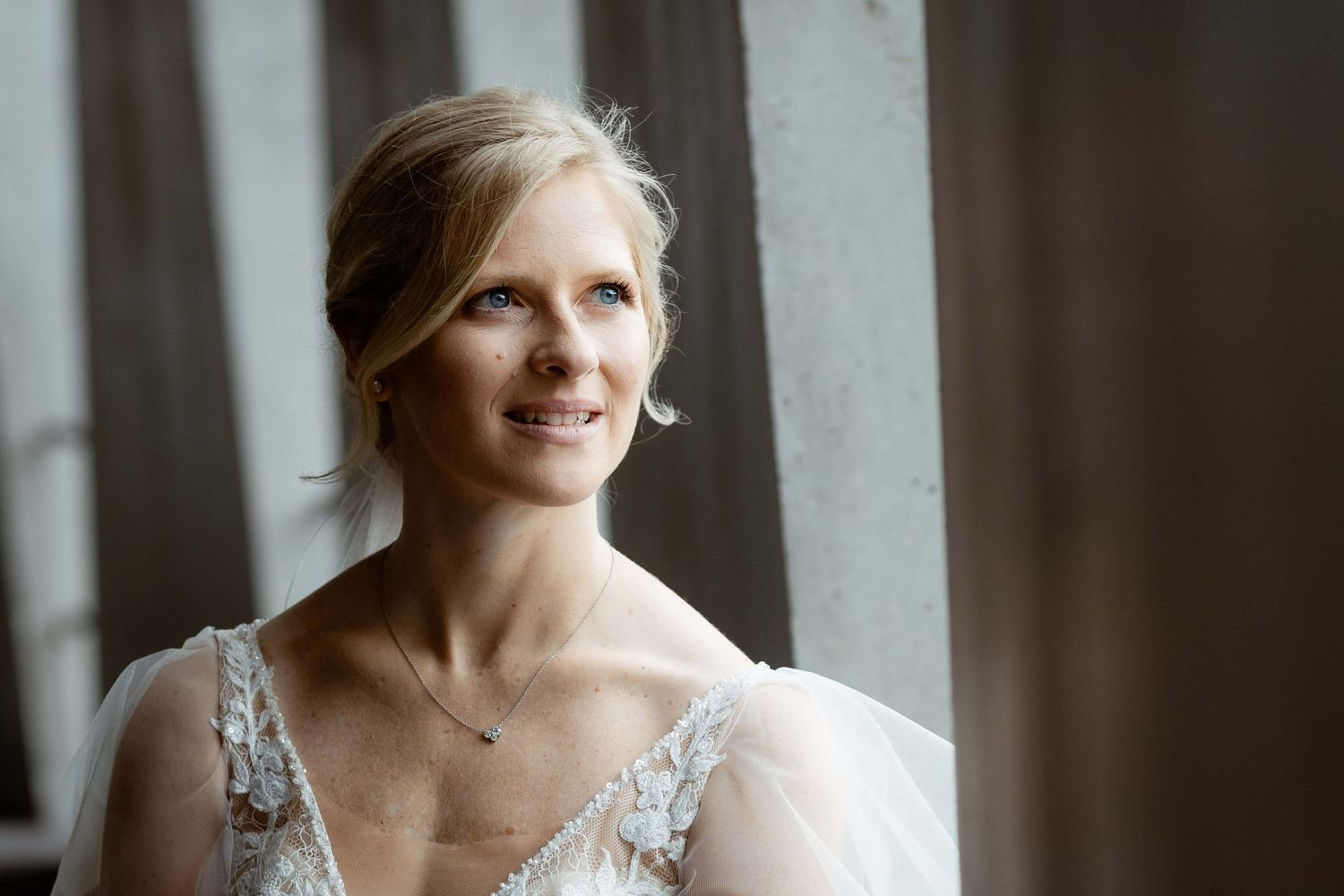 Smiling bride in lace wedding dress with delicate necklace gazes toward natural window light.