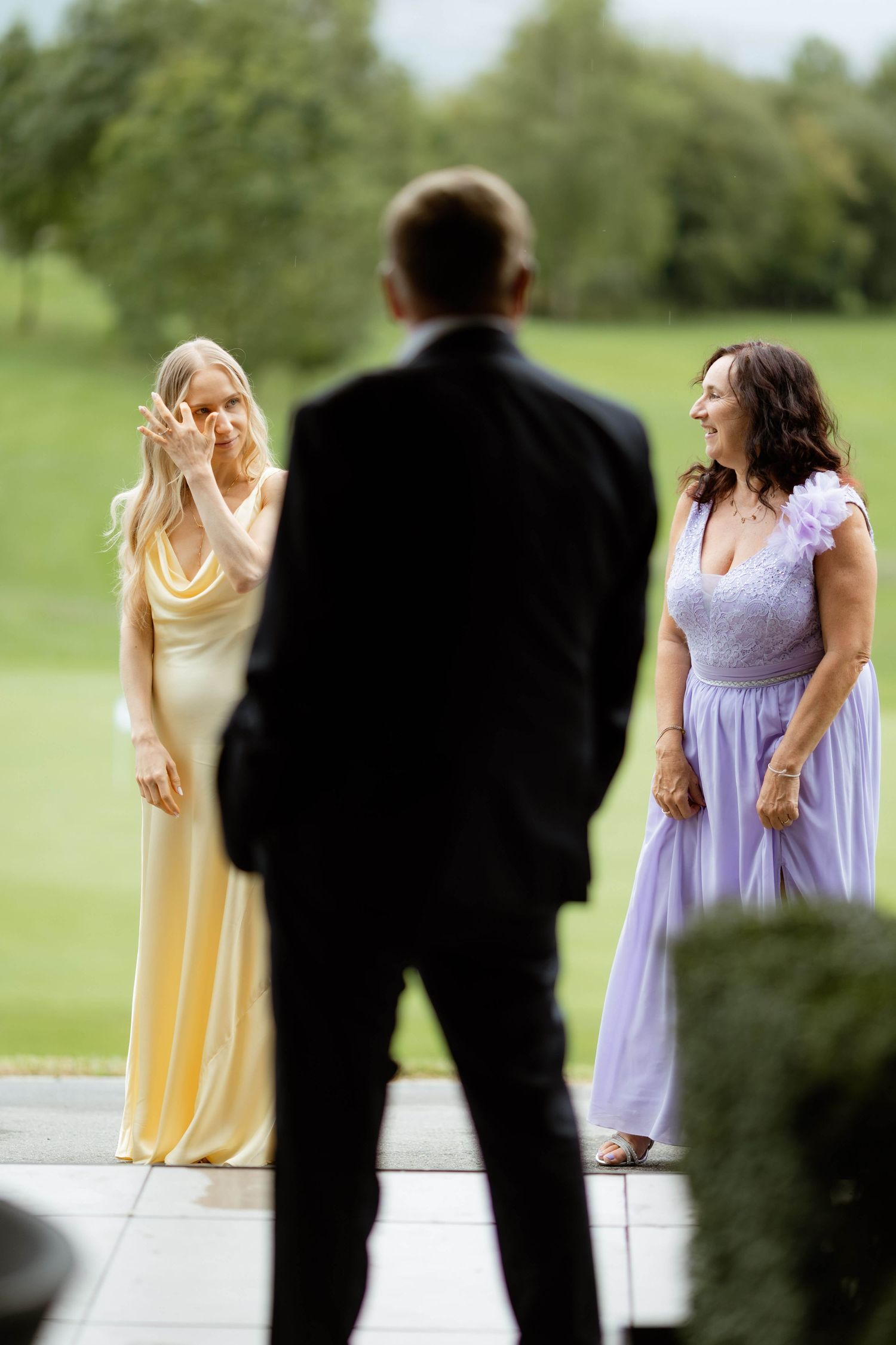 Two bridesmaids in yellow and lavender gowns react emotionally as a groom in a black suit approaches during an outdoor wedding.